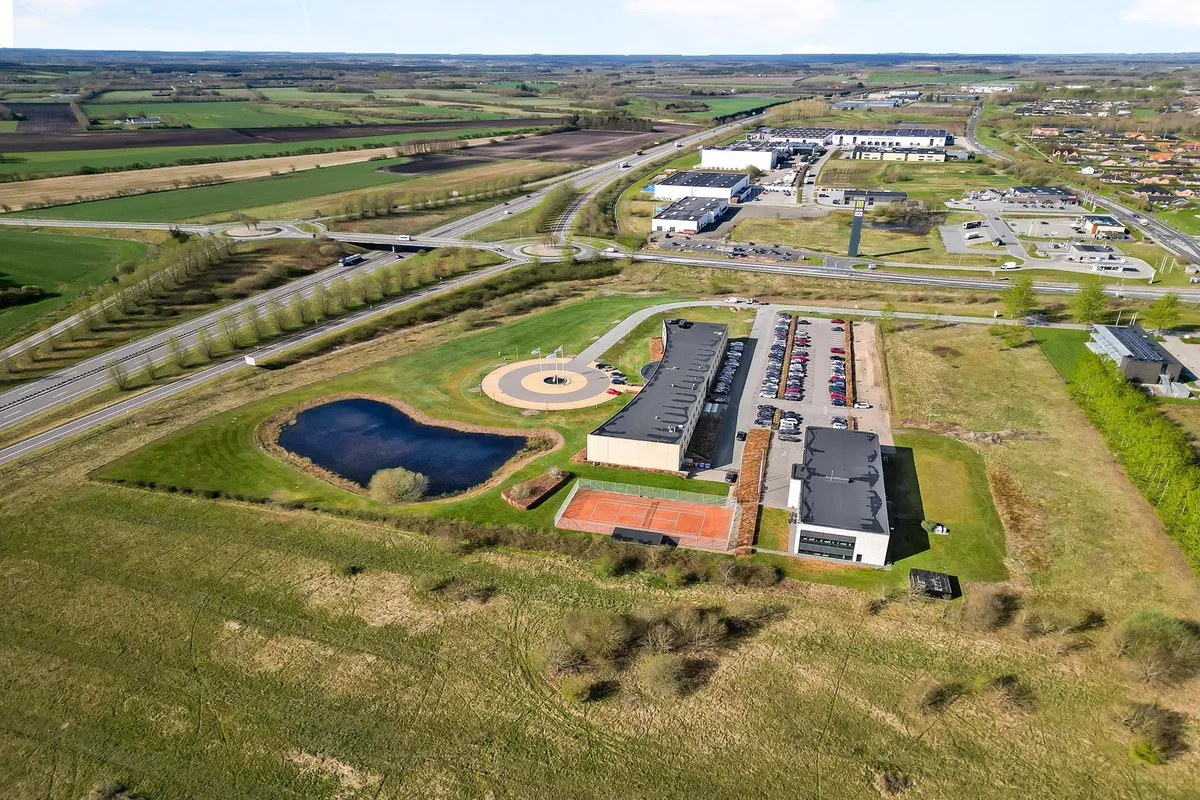 Bygning med parkeringsplads og tennisbane, omgivet af grønne marker og en sø. En bred hovedvej krydser området med flere