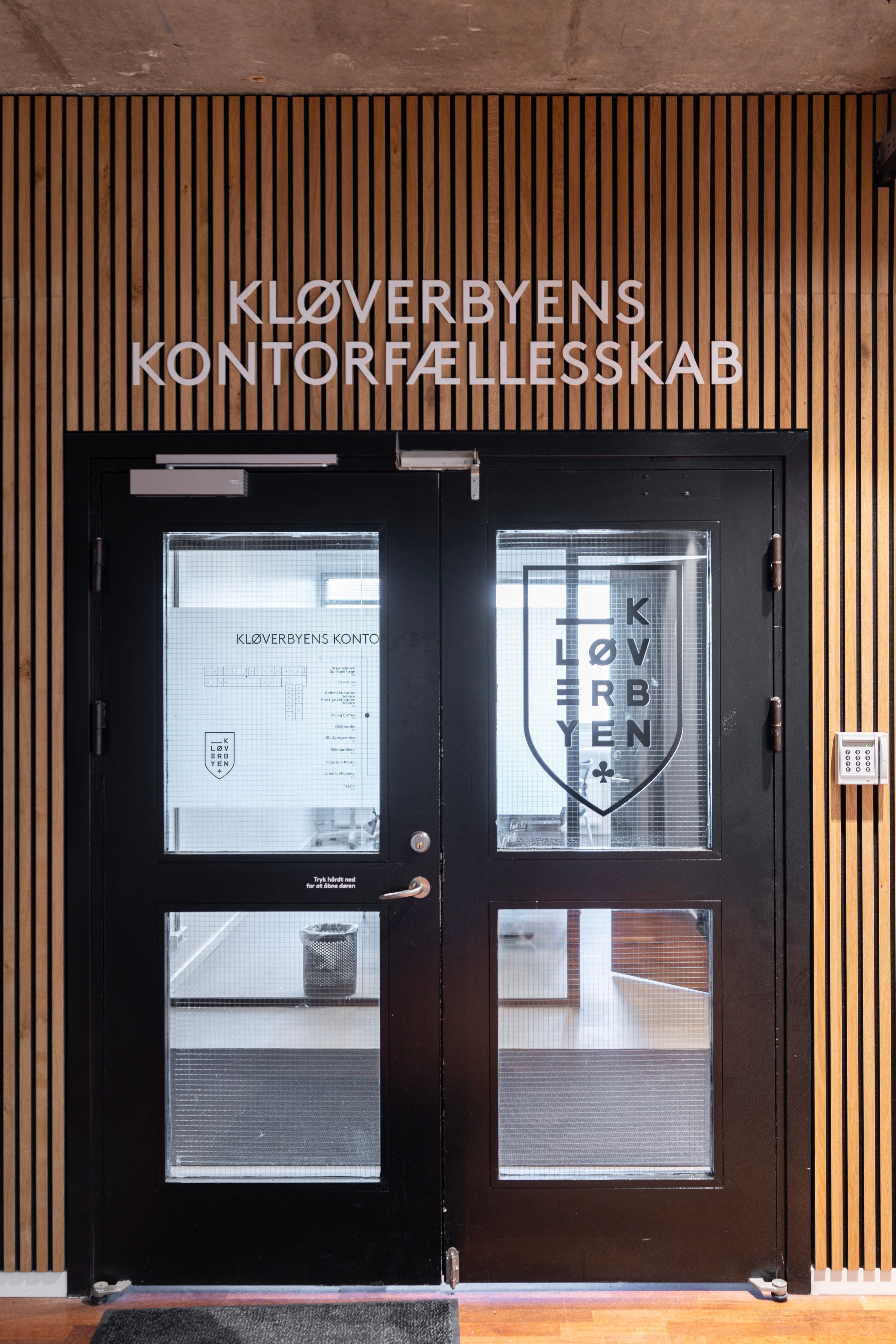 Sort glasdør med skrift Kloverbyens Kontorfaellesskab og logo med tekst Kloverbyen i sølv og sort