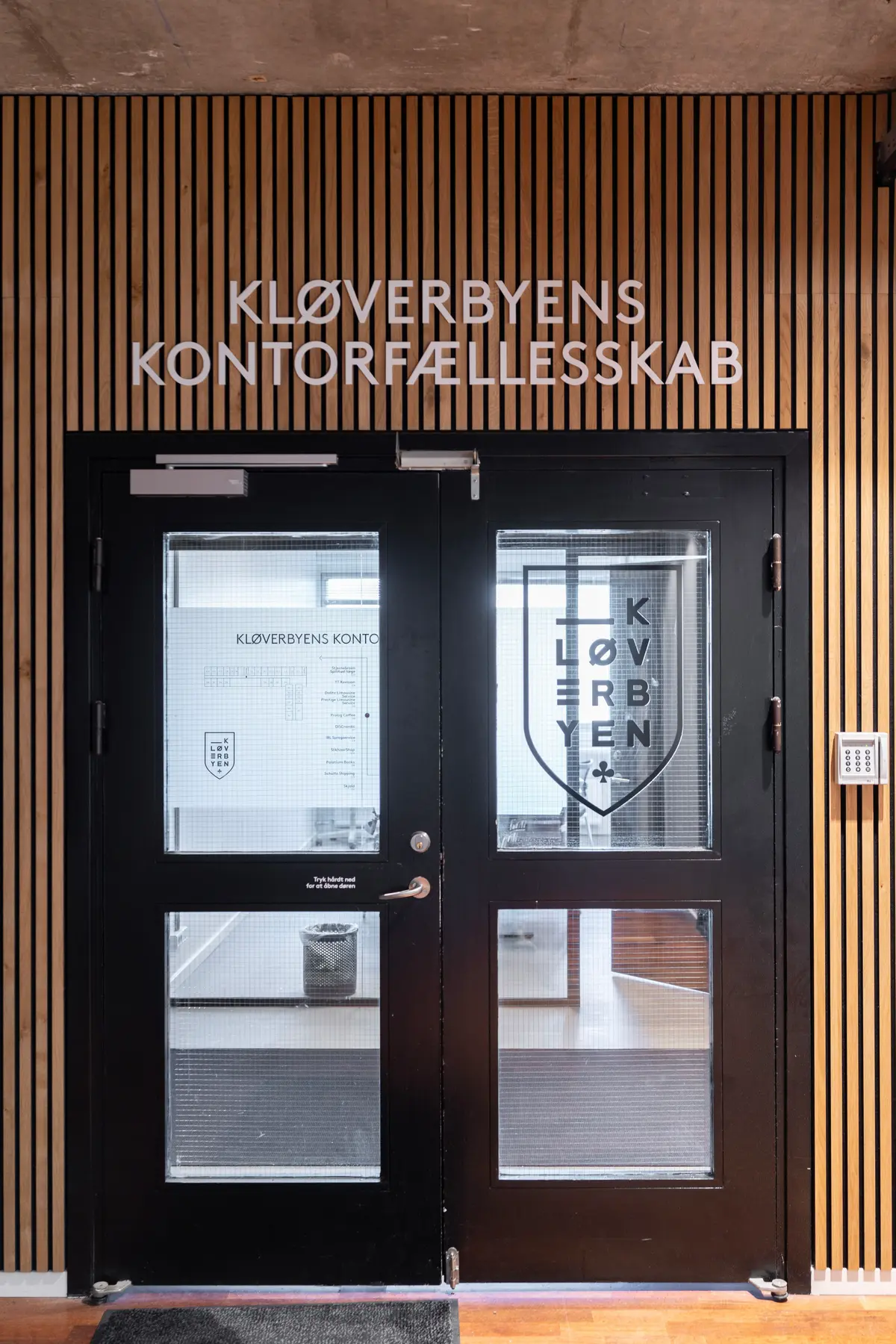 Sort glasdør med skrift Kloverbyens Kontorfaellesskab og logo med tekst Kloverbyen i sølv og sort