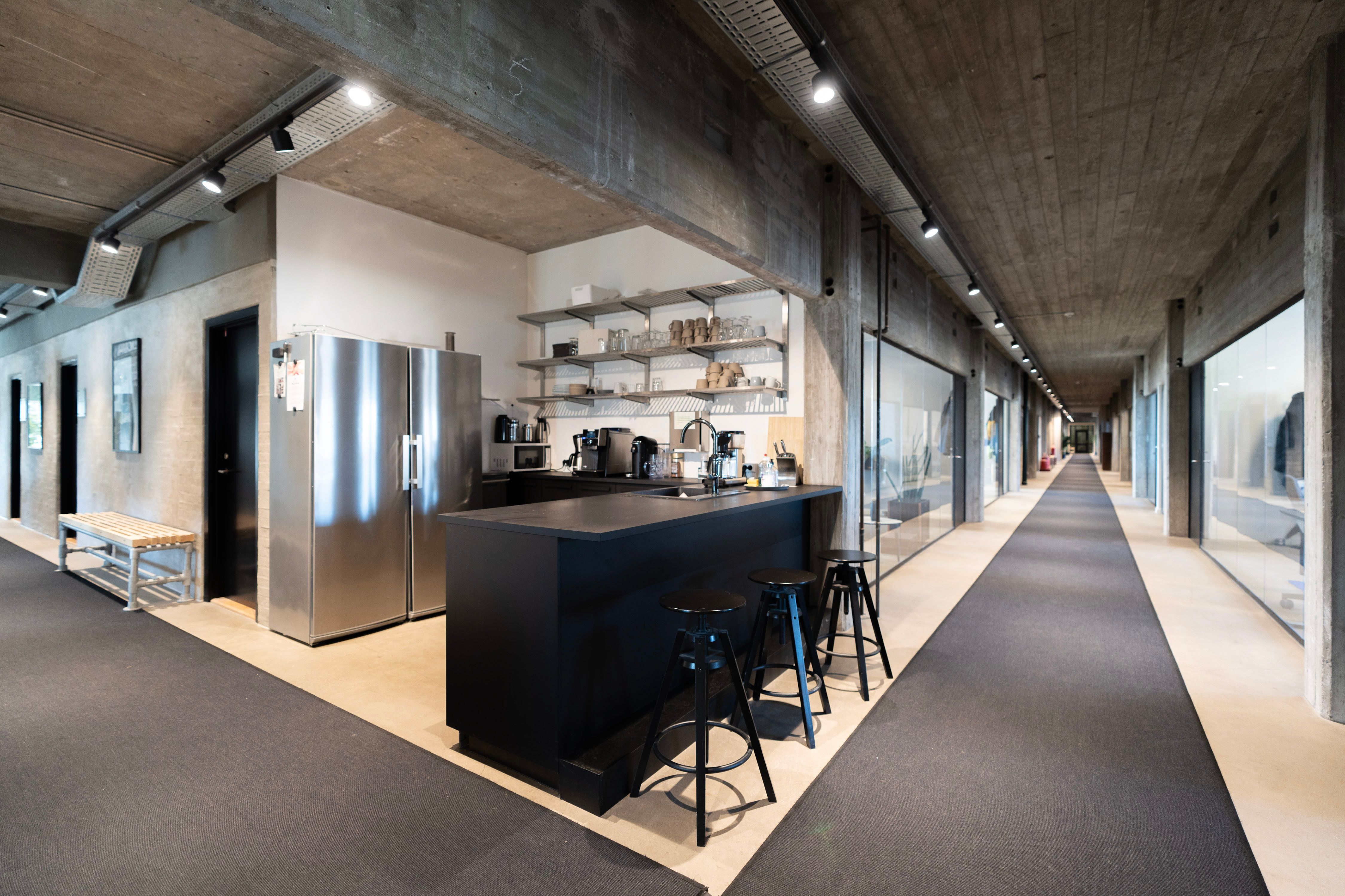 Kontorområde med lyse lofter og store glasruder, stolpe, køleskab og kogeapparater på bardisk