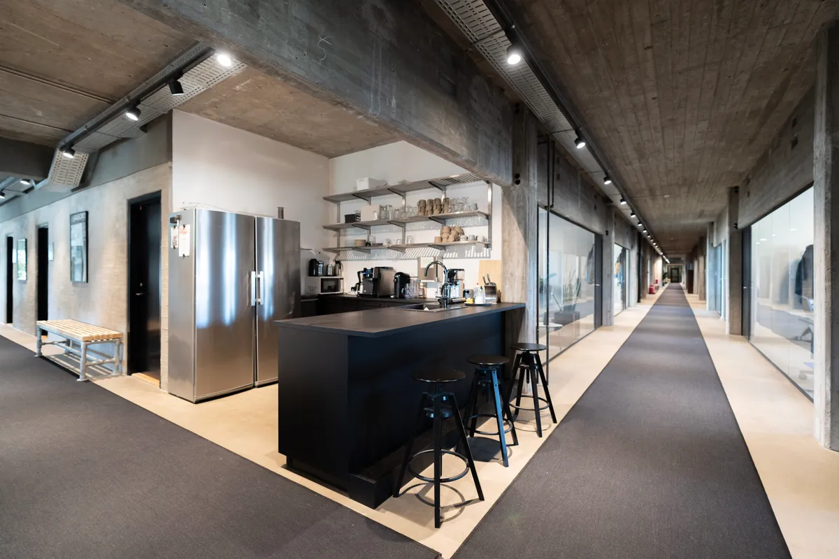 Kontorområde med lyse lofter og store glasruder, stolpe, køleskab og kogeapparater på bardisk