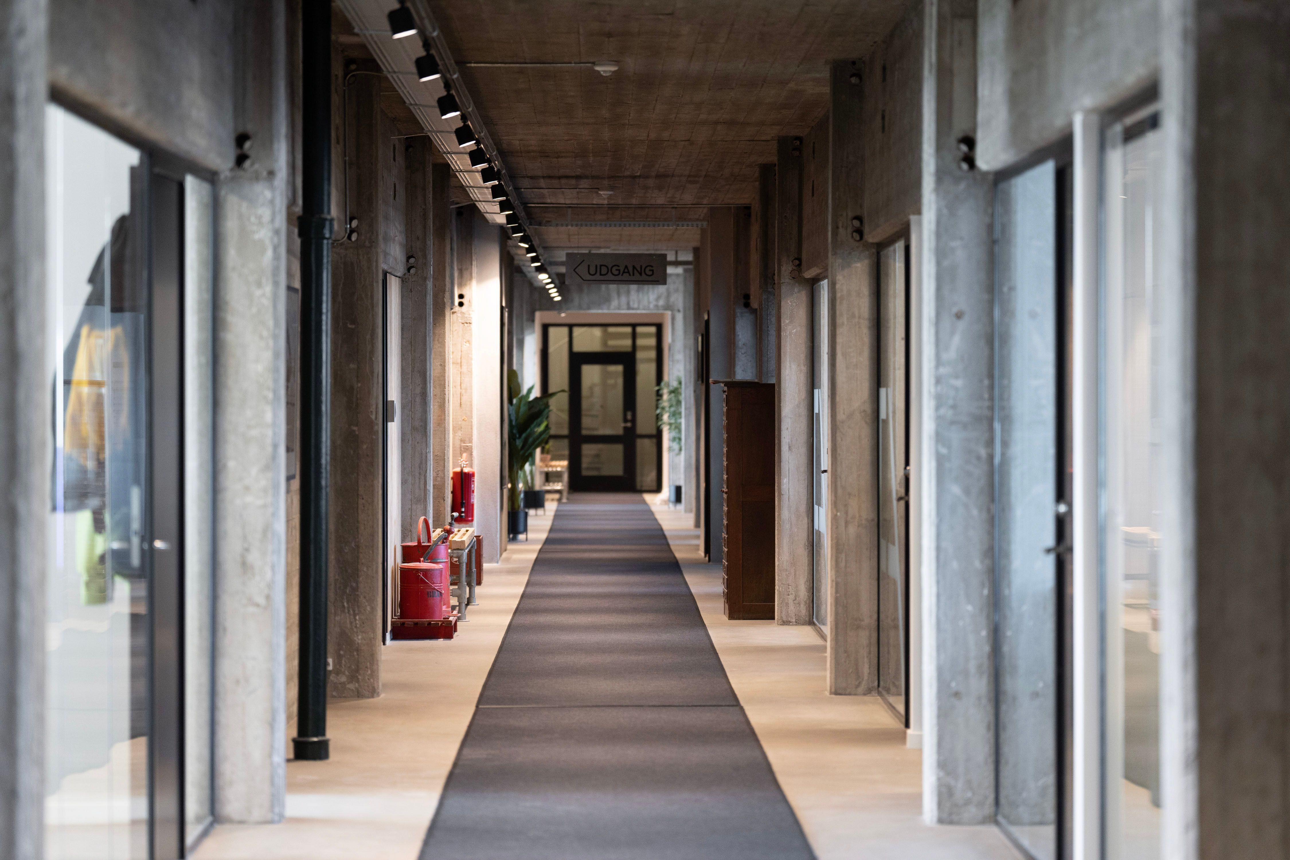 Kort og smal gangvej i et kontorfællesskab med glasvinduer, lysekroner og røde brandslukkere.