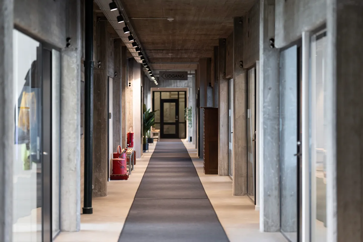 Kort og smal gangvej i et kontorfællesskab med glasvinduer, lysekroner og røde brandslukkere.