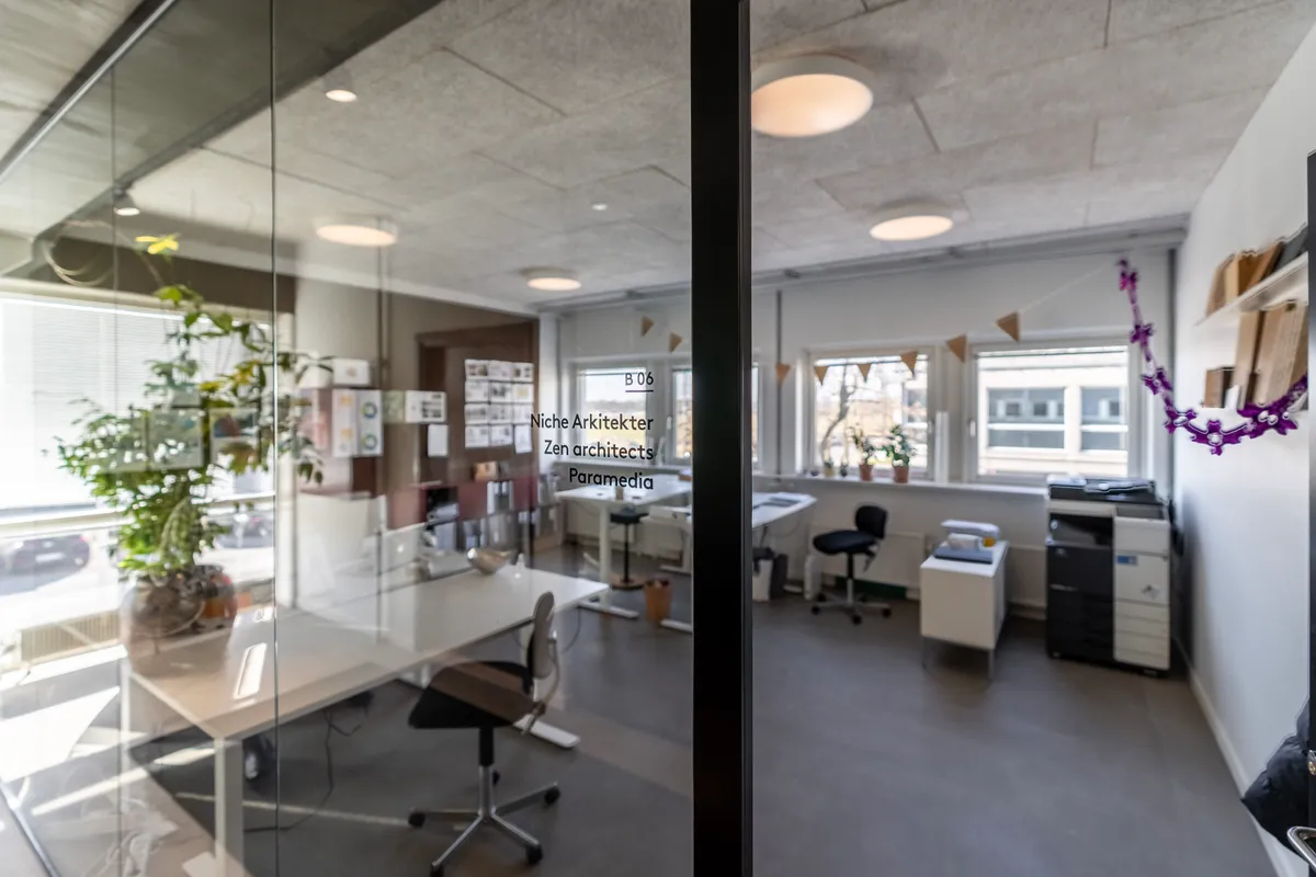 Kontor med store vinduer og grønne planter samt en skrivebord og en printer.