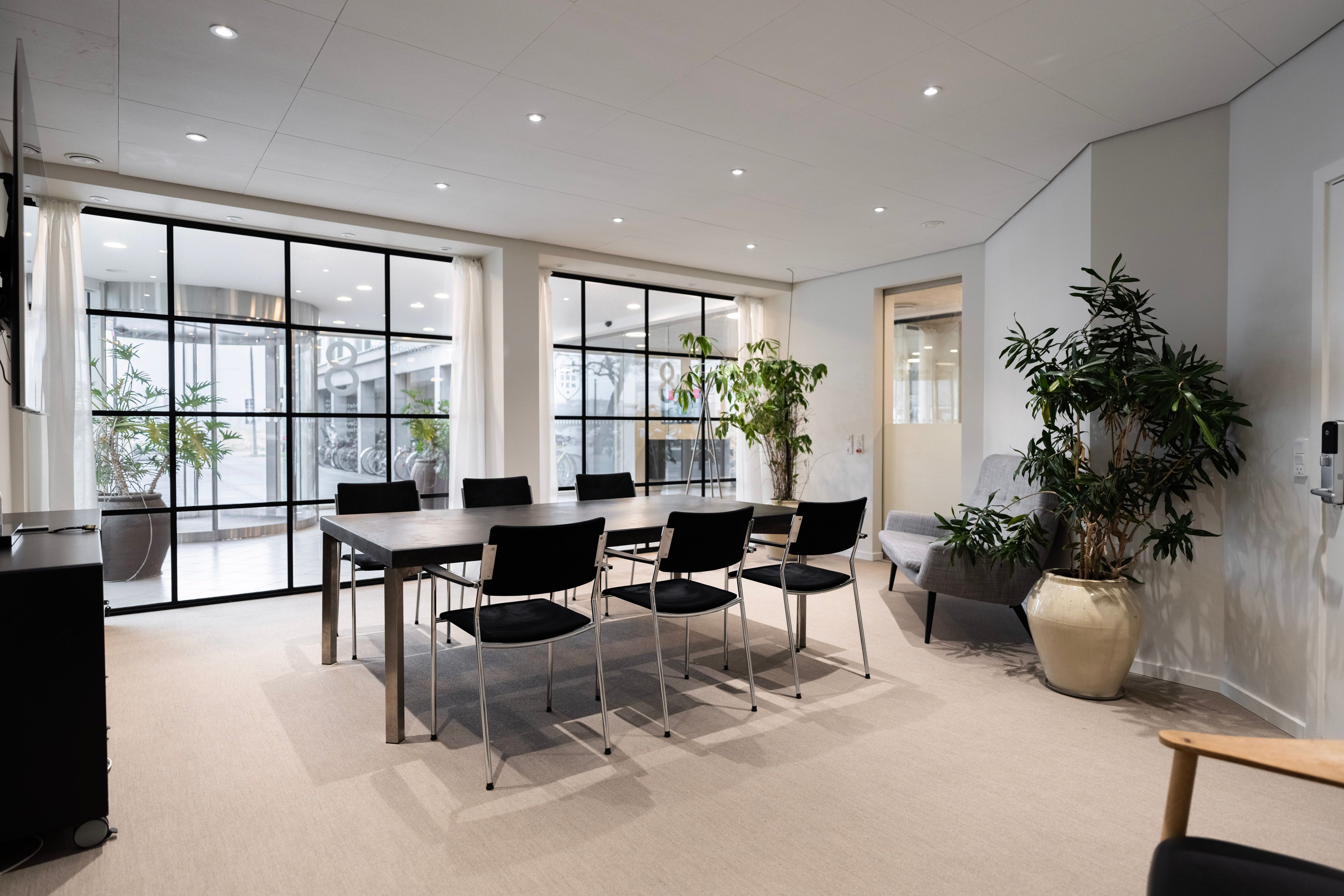 Kontor med lyse gulve, møbler og planter, en stor tabel og vinduer med store ruder