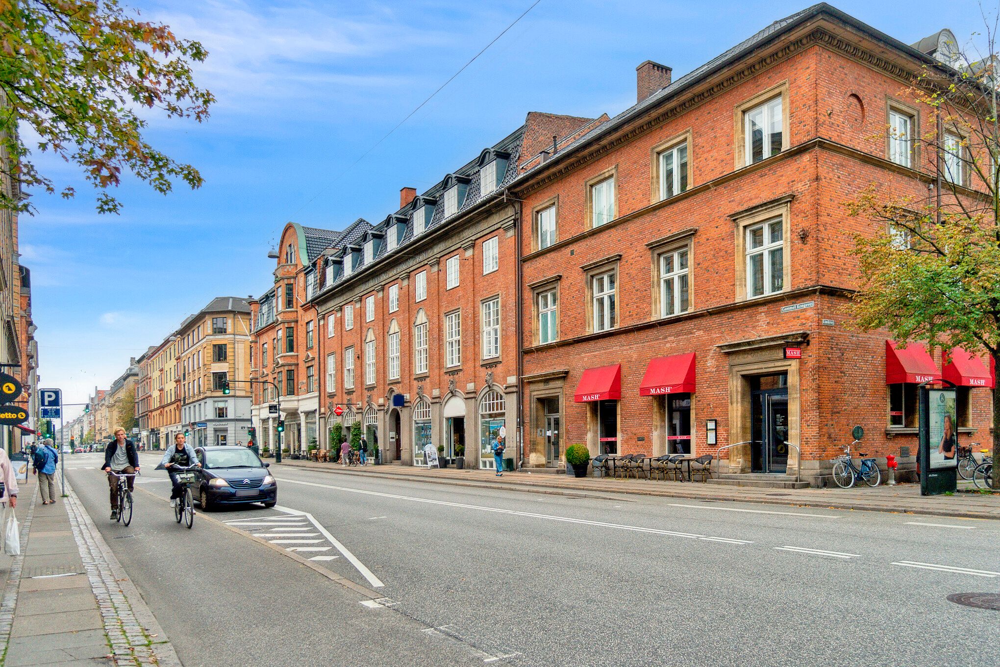 To cyklister kører langs Frederiksbergs hovedvej, og en bil passerer dem. På gadehjørnet er der en café og butikker med røde