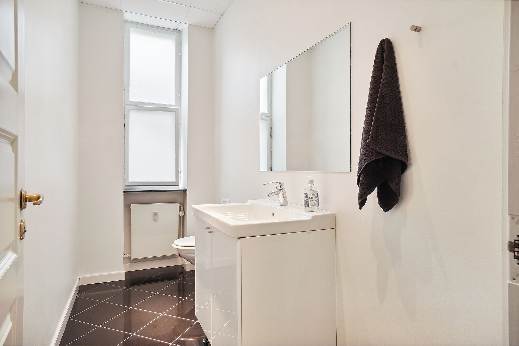 På dette billede ses et hvidt badeværelse med toilet, spejl, håndklæde og håndvask med vandhaner og sæbeflaske.