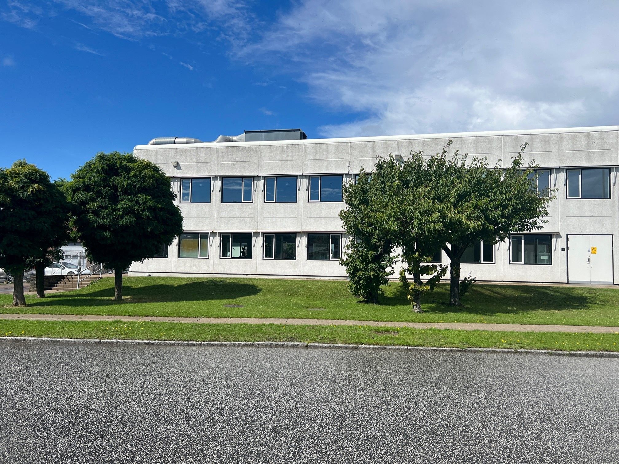 Et kontorblok med flere vinduer og en hvid dør ligger ved siden af en asfaltvej med grønne græsplæner og to små træer