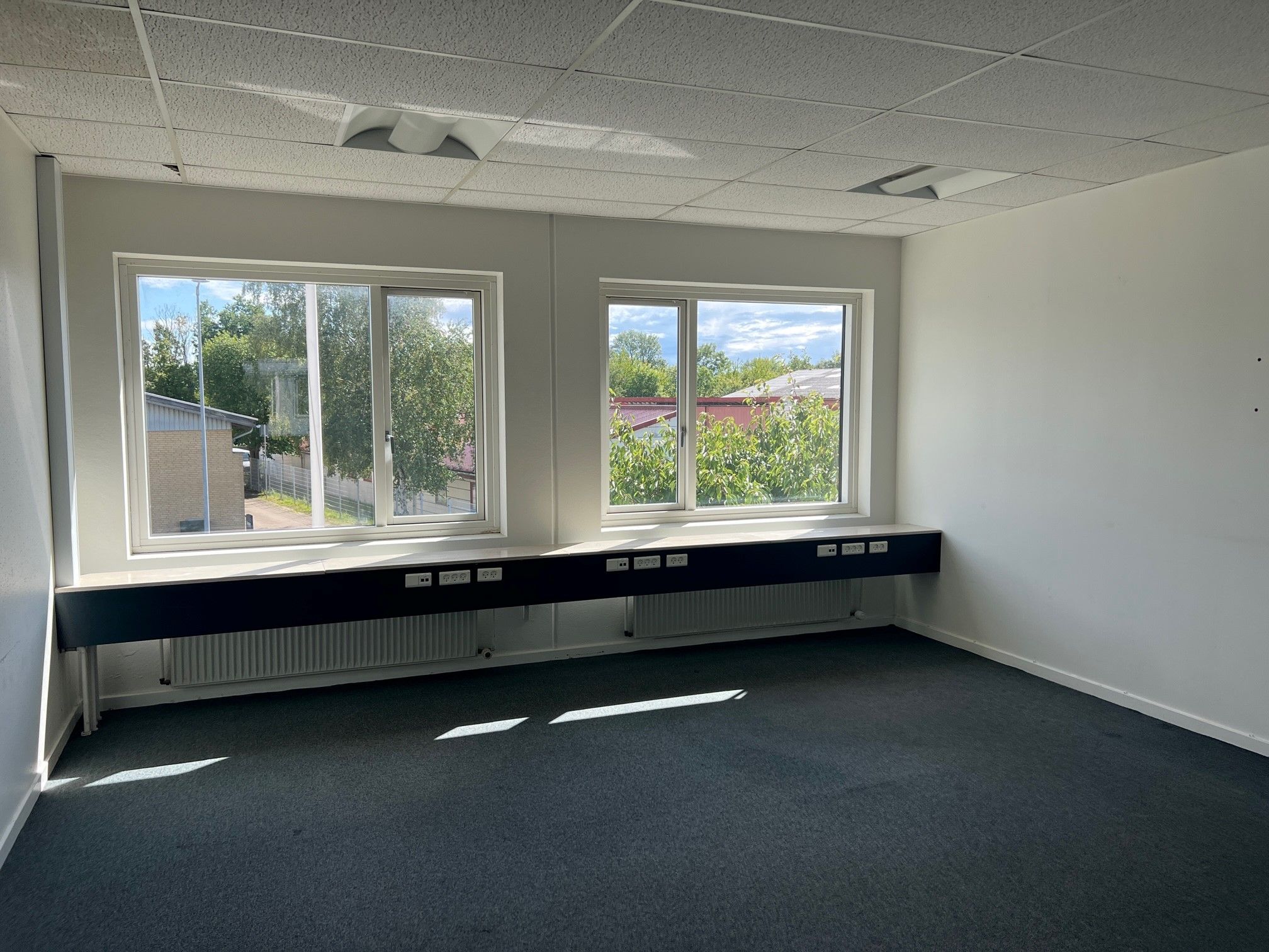 Et tomt kontor med hvide vægge, gulv og loft, to store vinduer og et langt sort skrankebord med elstik