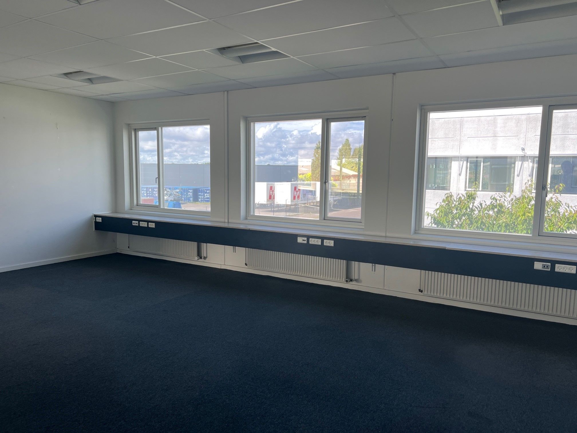 Et stort, tomt kontorrum med vinduer og radiatorer under vinduet samt mørkegulve og hvide vægge
