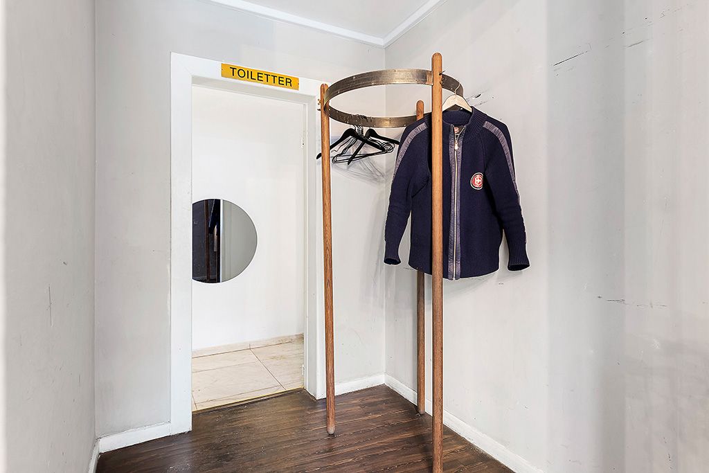 En slank tøjstang med to hængere og en hvid trøje i et lille, hvidmalet rum med et trægulv og et toiletskilt