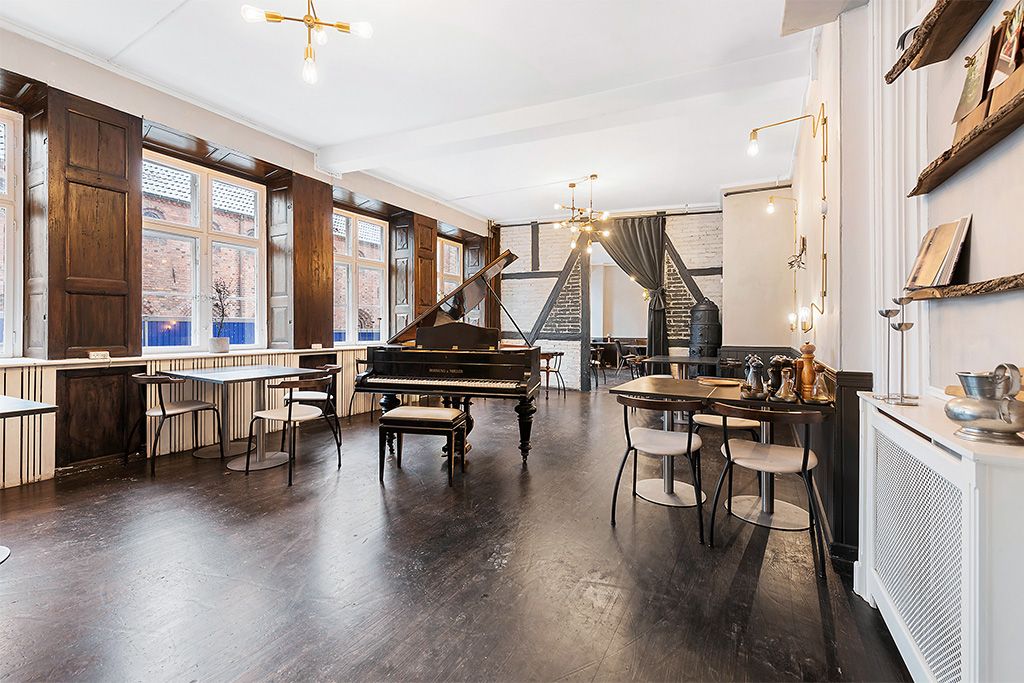 En stor, lys stue med et stolpepiano, lysekroner, hvide vægge, trægulve og møbler