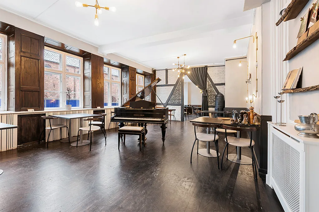 En stor, lys stue med et stolpepiano, lysekroner, hvide vægge, trægulve og møbler