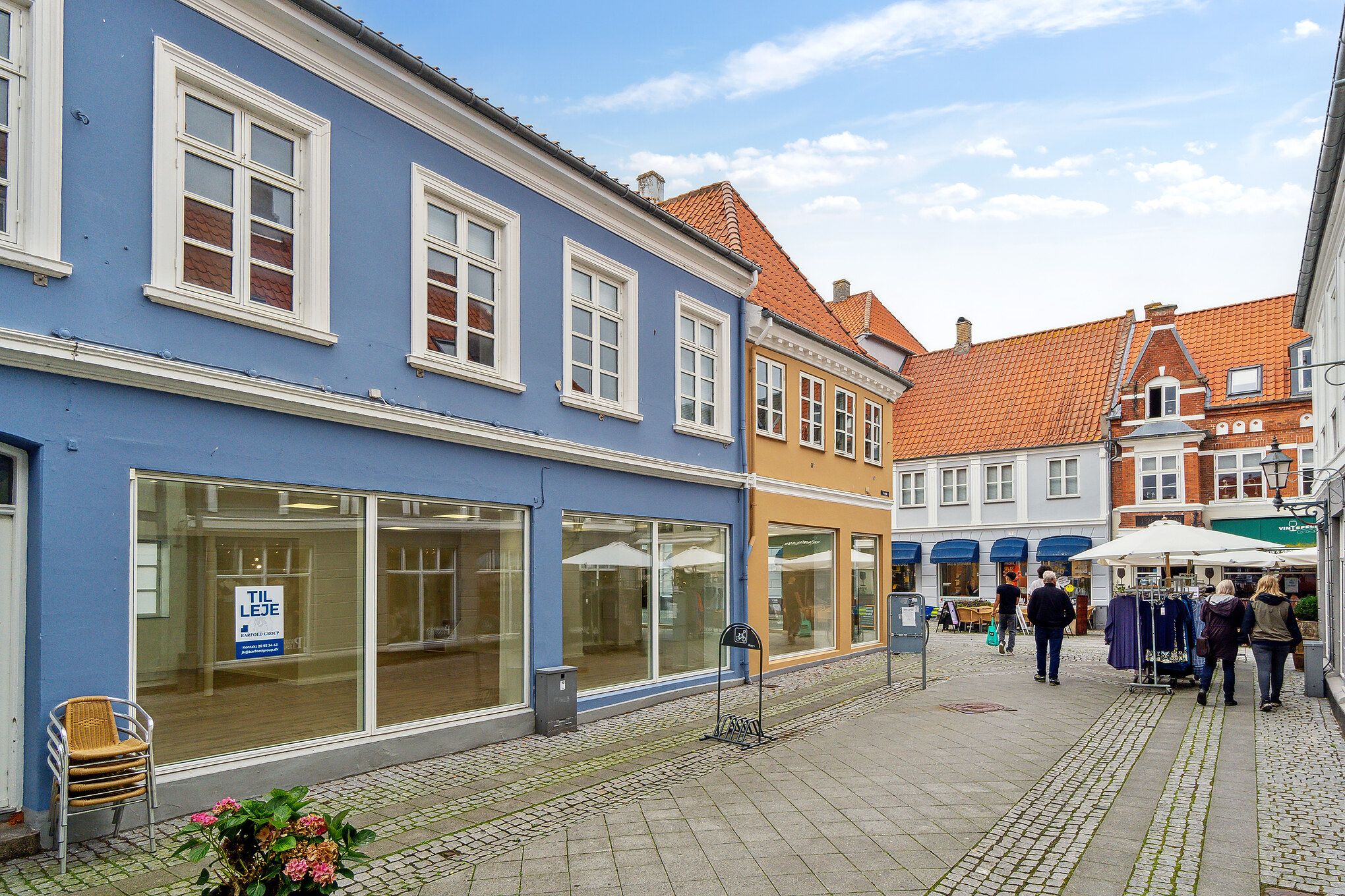 Købmandsgade i Nyborg med forretningslokaler, hvor en butik er til leje, og tre personer går forbi en klædeskole med tøj i