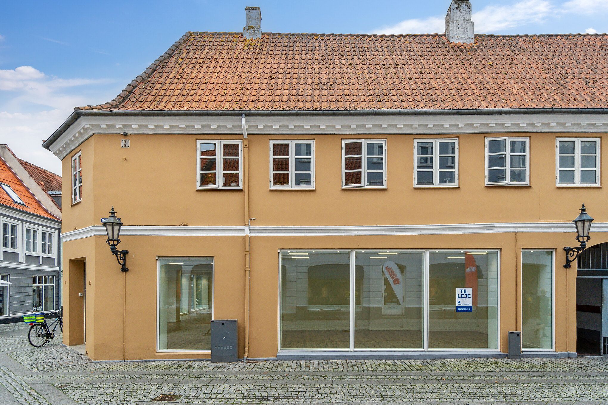 En butik med en stor facade og flere vinduer i Nyborg med en til leje skilt og en cykel uden for