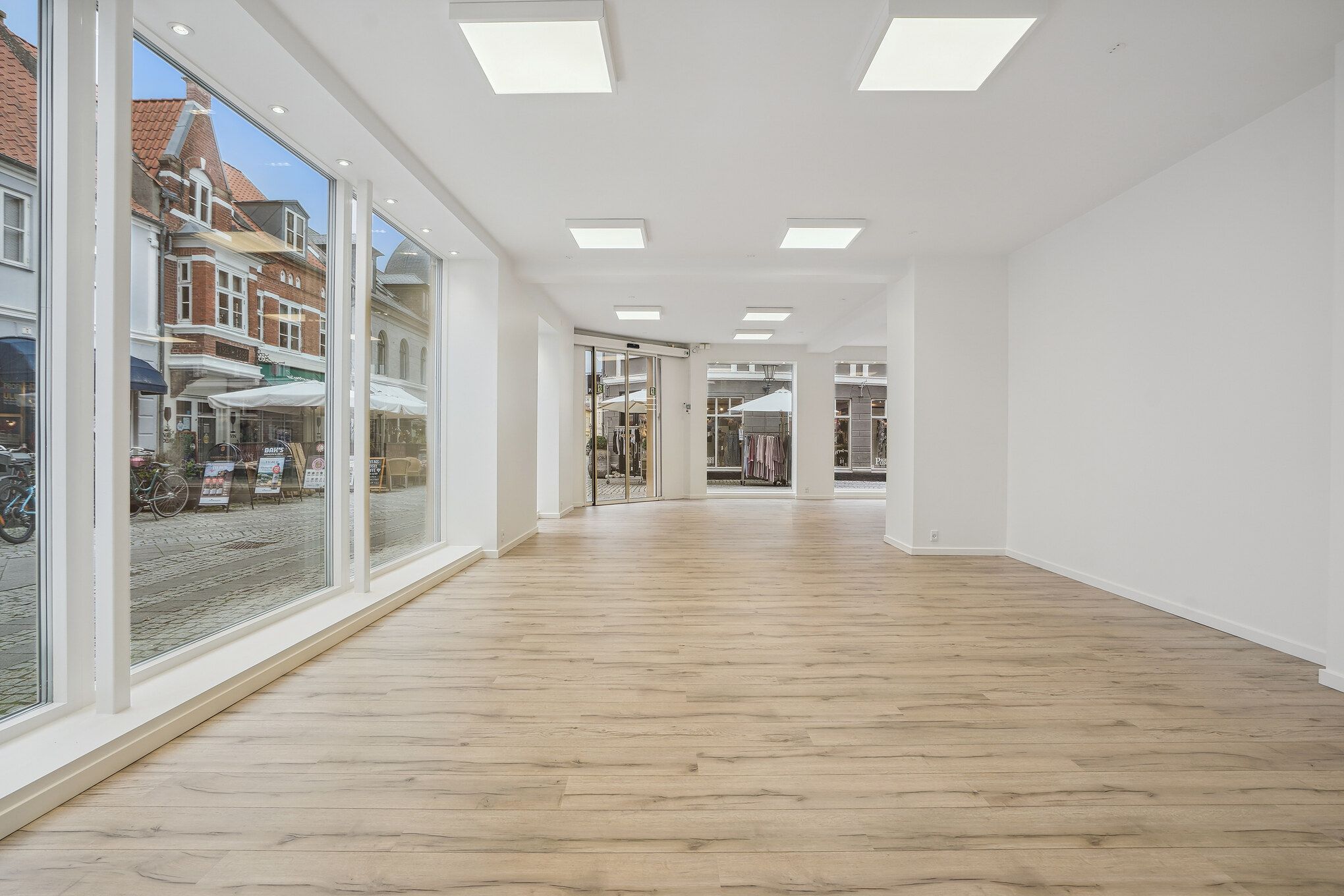 Tomme lokaler med høje glasvinduer og lyse loftslamper, hvide vægge og lysegul trægulv og en åben dør med udgang til