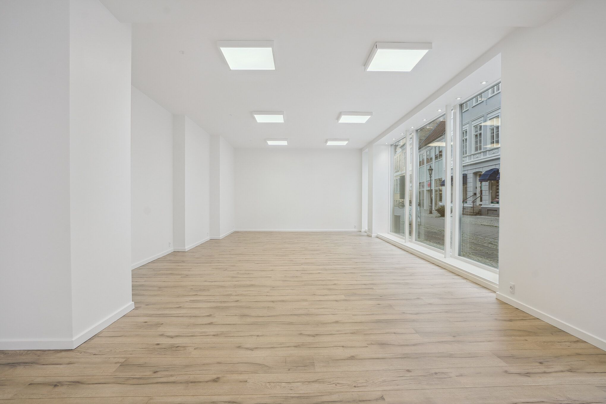 En moderne, tom og velrenoveret butik med lyse loftslampe, hvide vægge, store glasvinduer og massiv trægulv, der vender mod