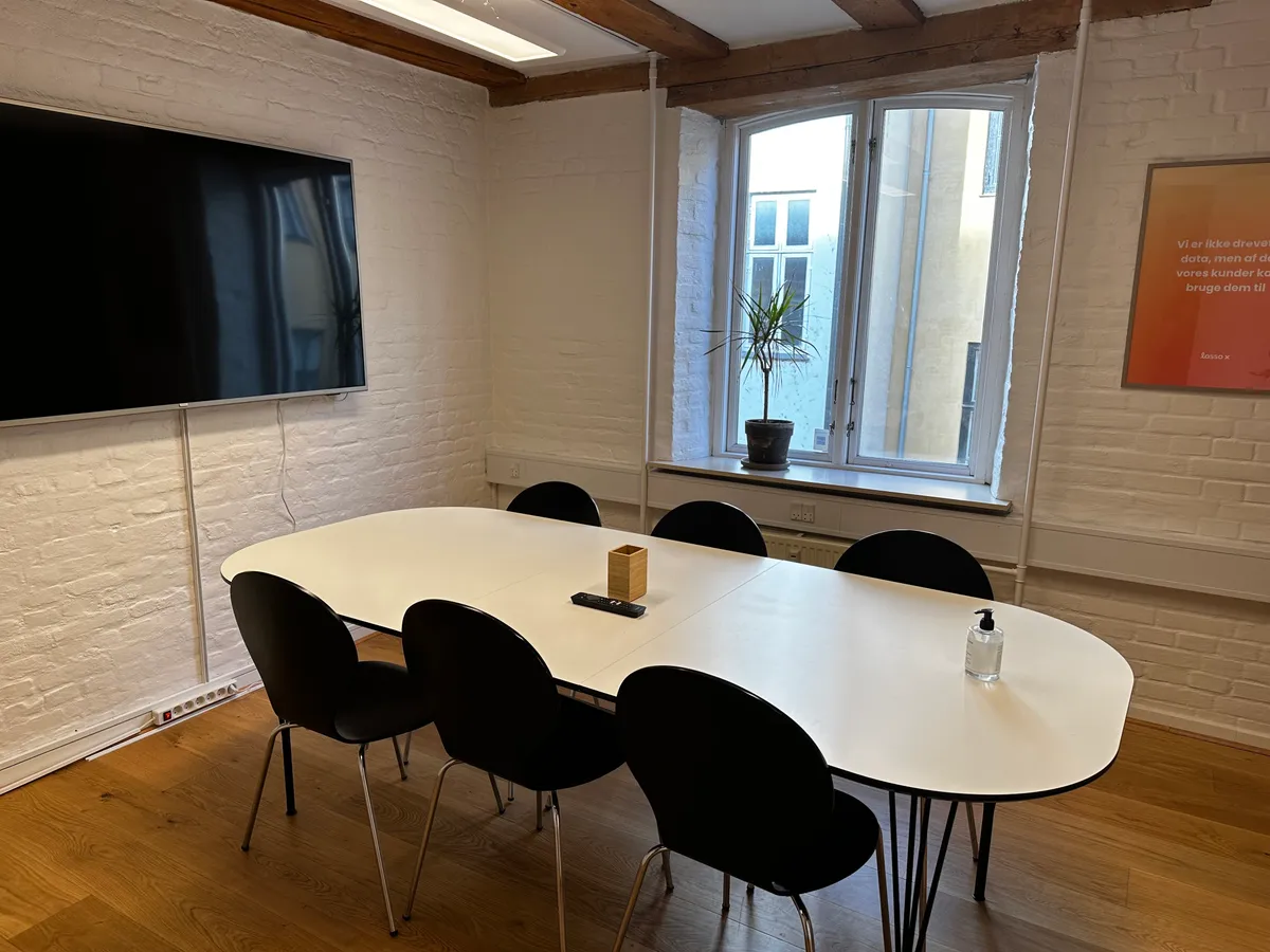 En mødelokale med en stor rund bord og stolpe. Der er en plante i vindueskarmen og en tv-skærm monteret på væggen.