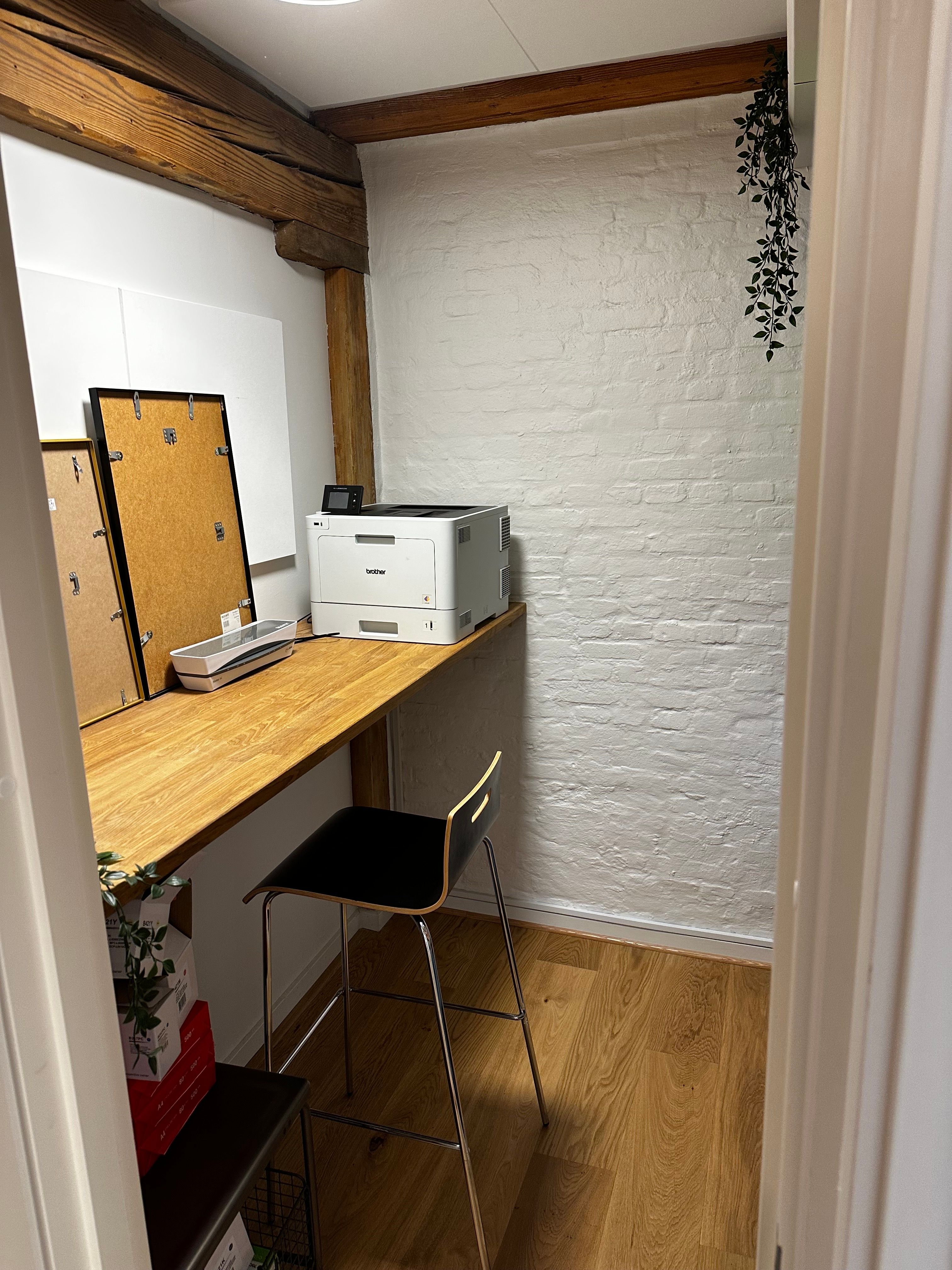 En lille kontor med en høj skrivebord, en Brother laserprinter og en stol foran en hvid mur og loft med et grønt hængende