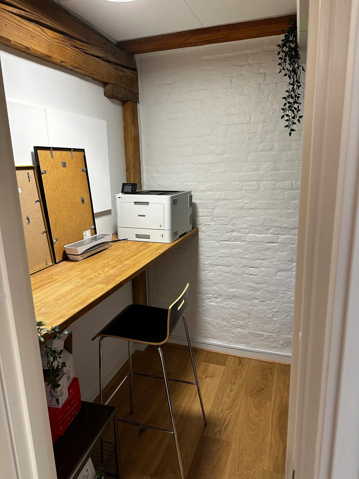 En lille kontor med en høj skrivebord, en Brother laserprinter og en stol foran en hvid mur og loft med et grønt hængende