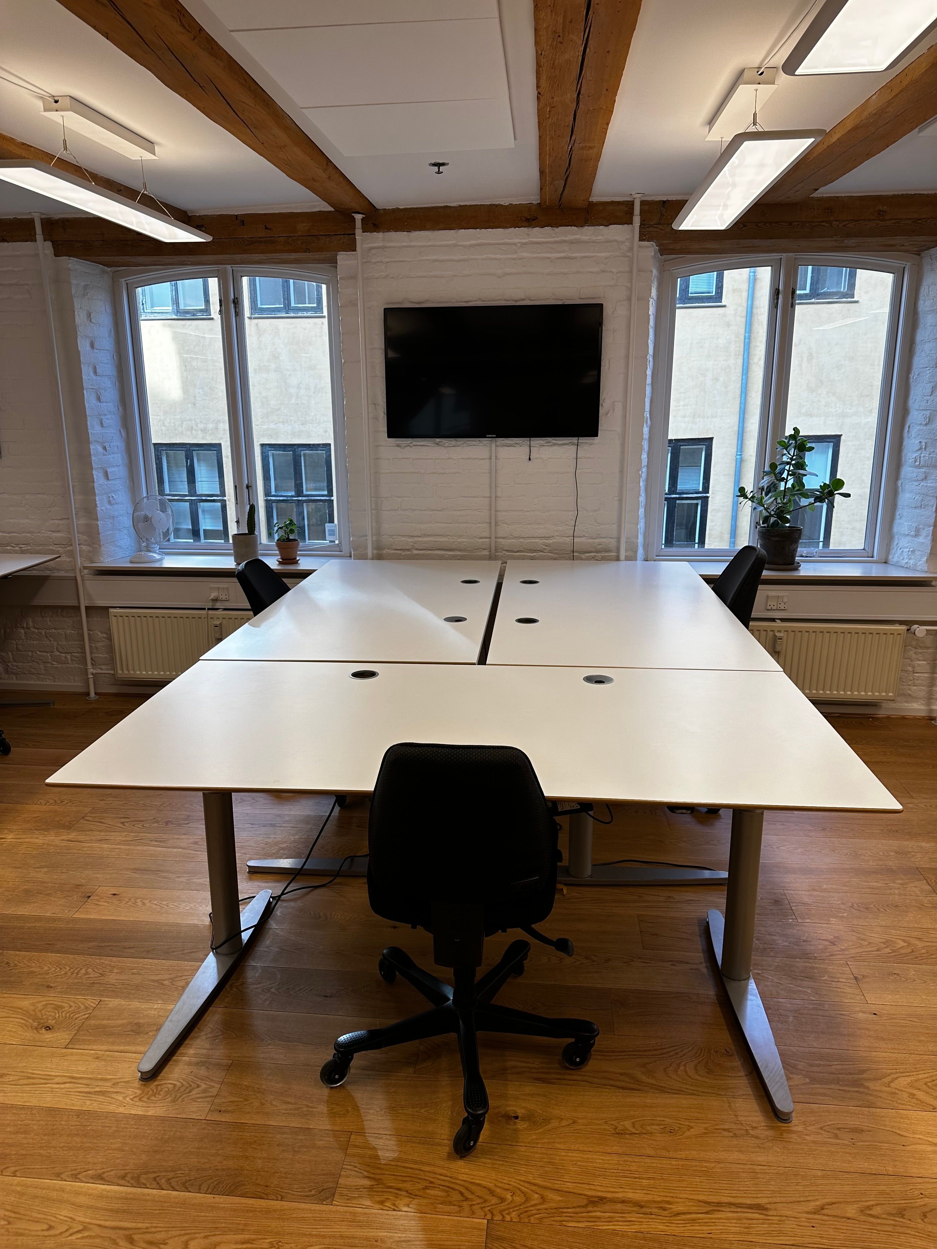Kontor med to hvide bord og fire møbler, hvor de to forreste er sorte og de to bageste er hvide, med en tv-skærm, lysekroner