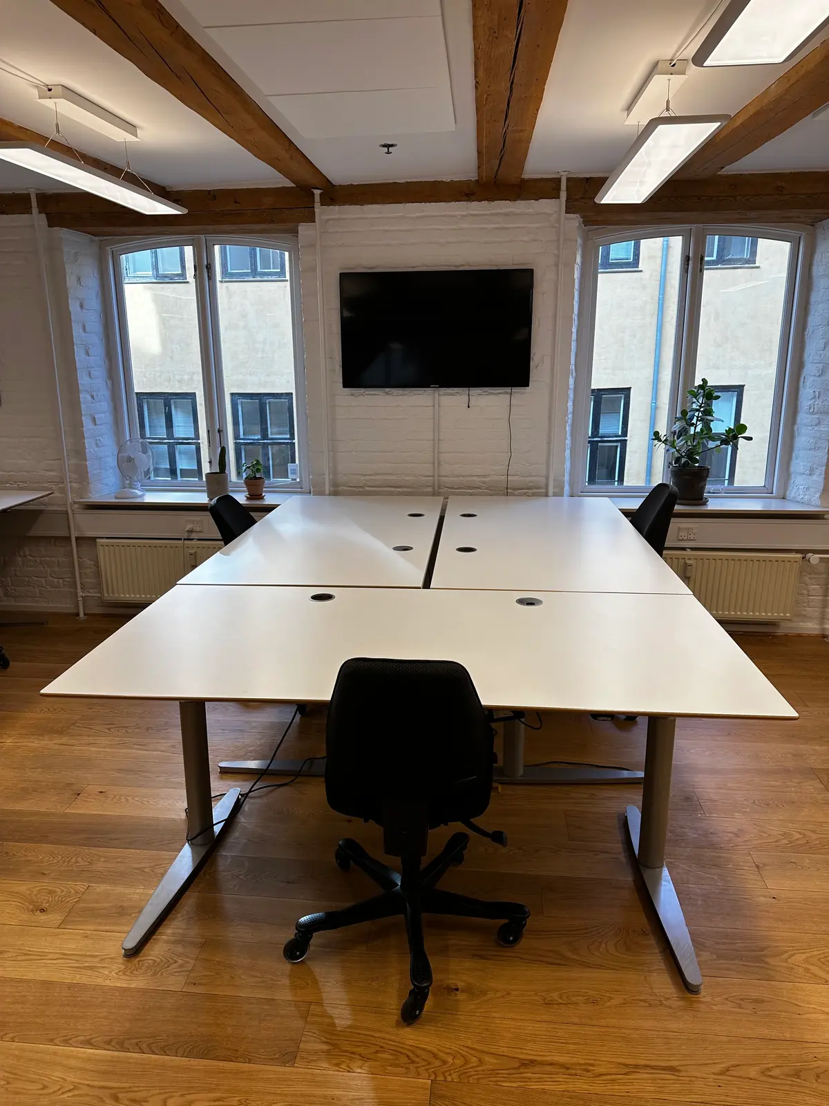 Kontor med to hvide bord og fire møbler, hvor de to forreste er sorte og de to bageste er hvide, med en tv-skærm, lysekroner