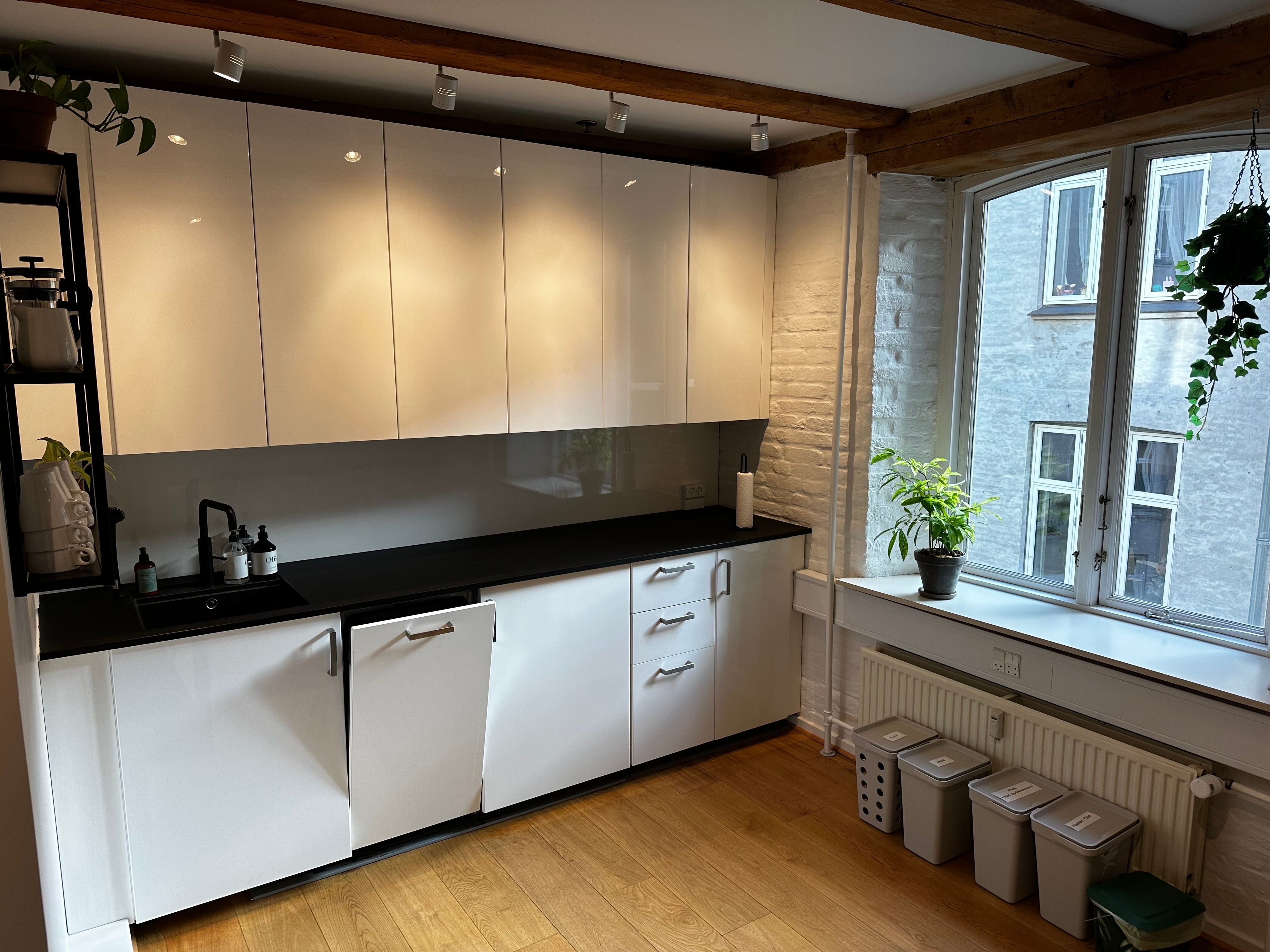 Et moderne køkken med hvide skabe, sort bordplade, og lyse pærelamper. Køkkenbordet har en kaffemaskine og et glas, og der