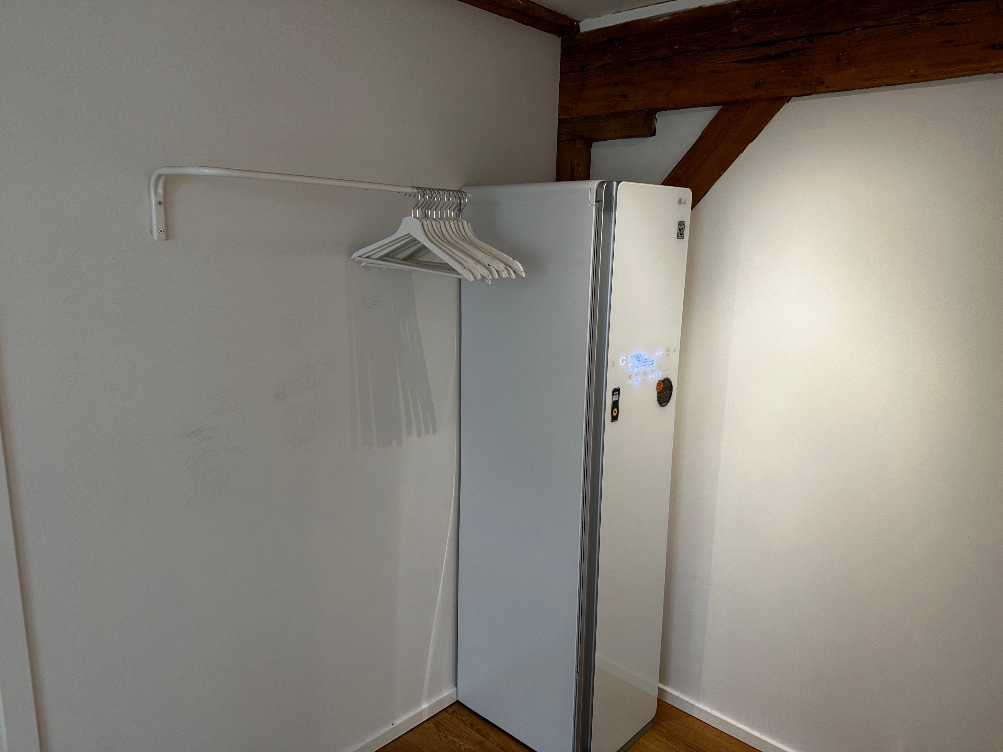 LG køleskab og hængehylde med hvide træhængere står i et hjørne med hvide vægge og træloft