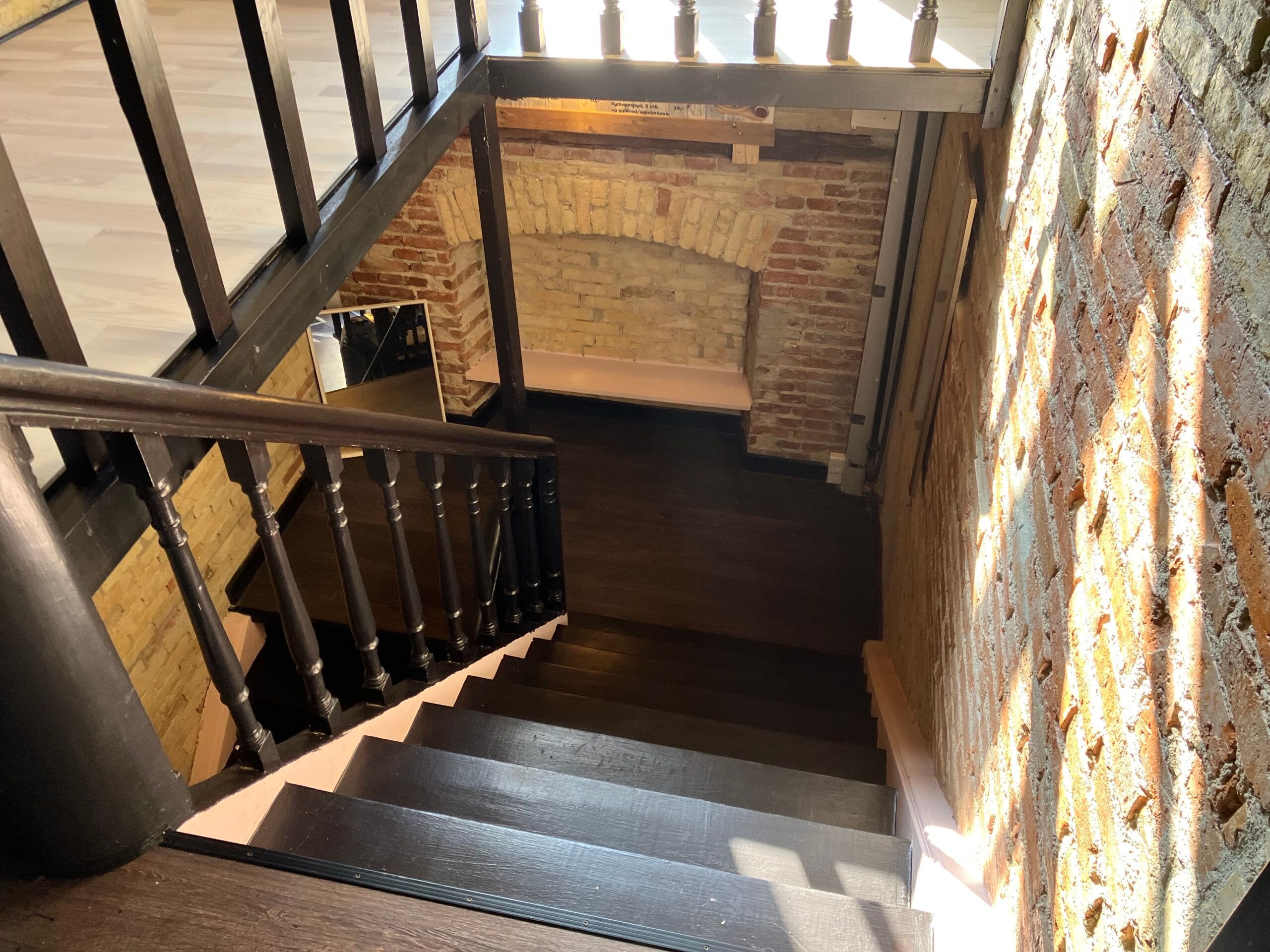 En trappe med mørkt træ og lyse håndtag, der fører ned til et rum med bjælkeloft og murstensvægge