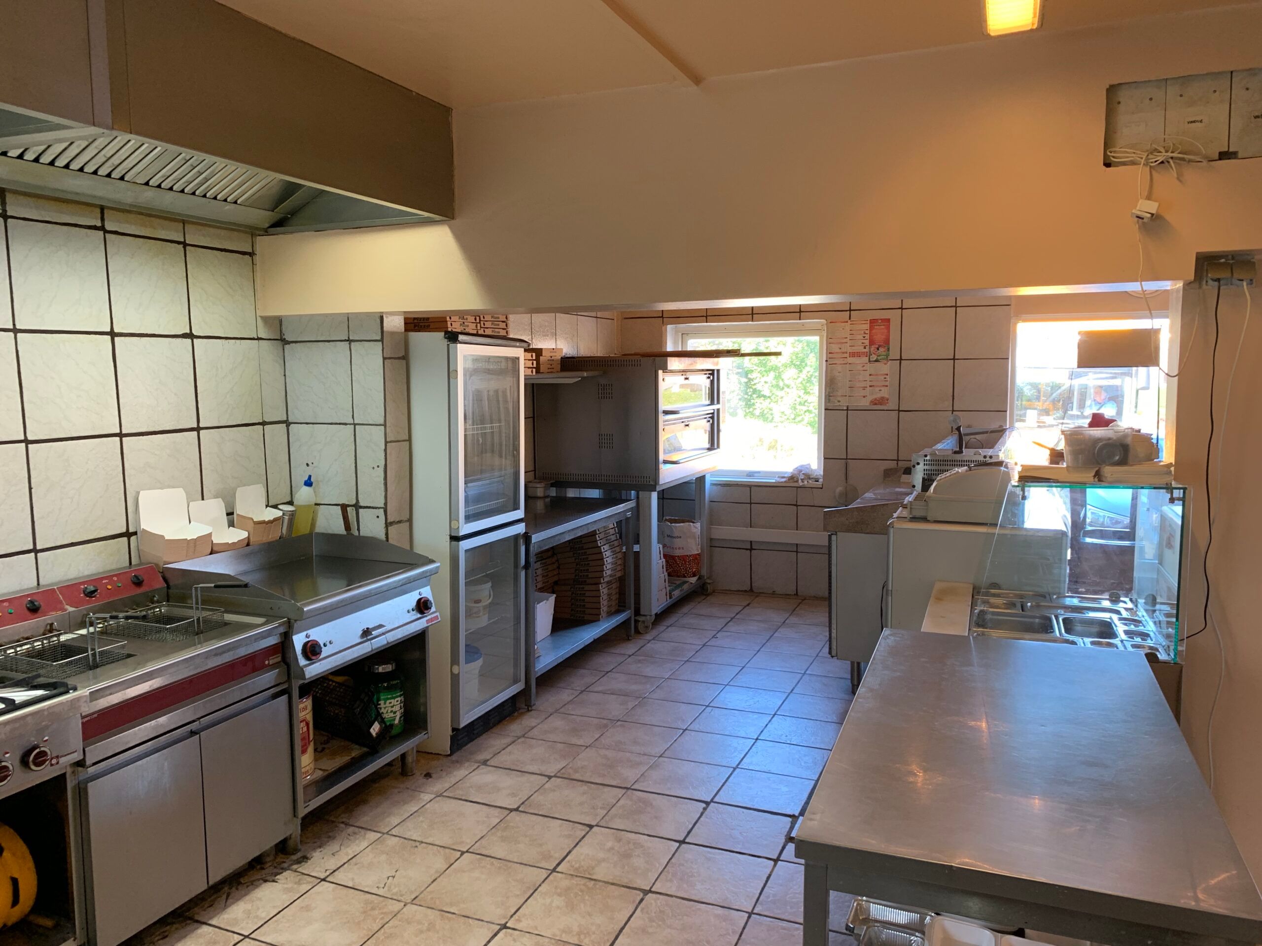 Rummelig køkken med stålborde, ovne, køleskabe, en vinduesåbning og en ventileringshatte