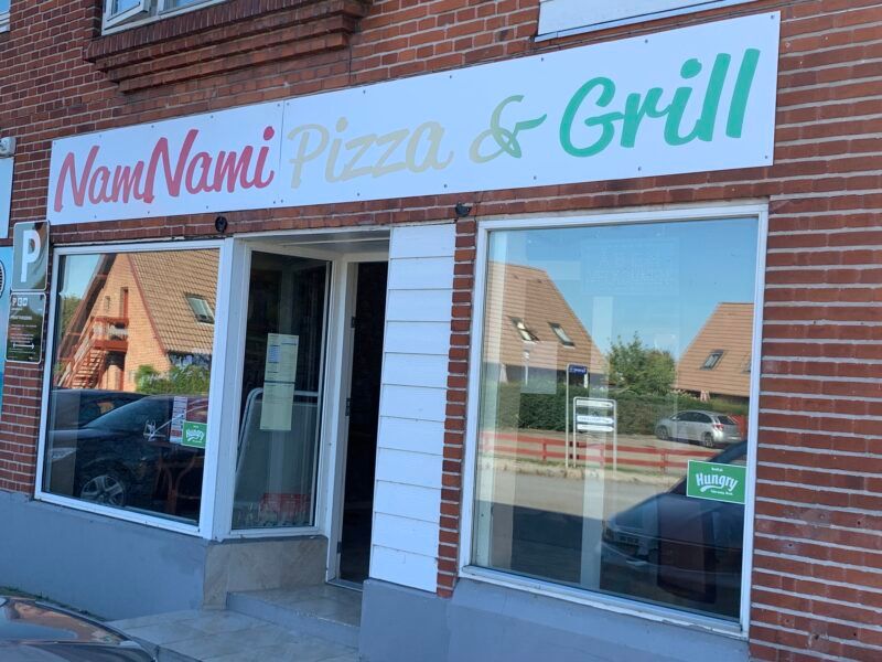 Pizza og grill restaurant med en hvid facade og skrift. Navnet er NamNami Pizza & Grill. Der er en åben dør og vinduer med