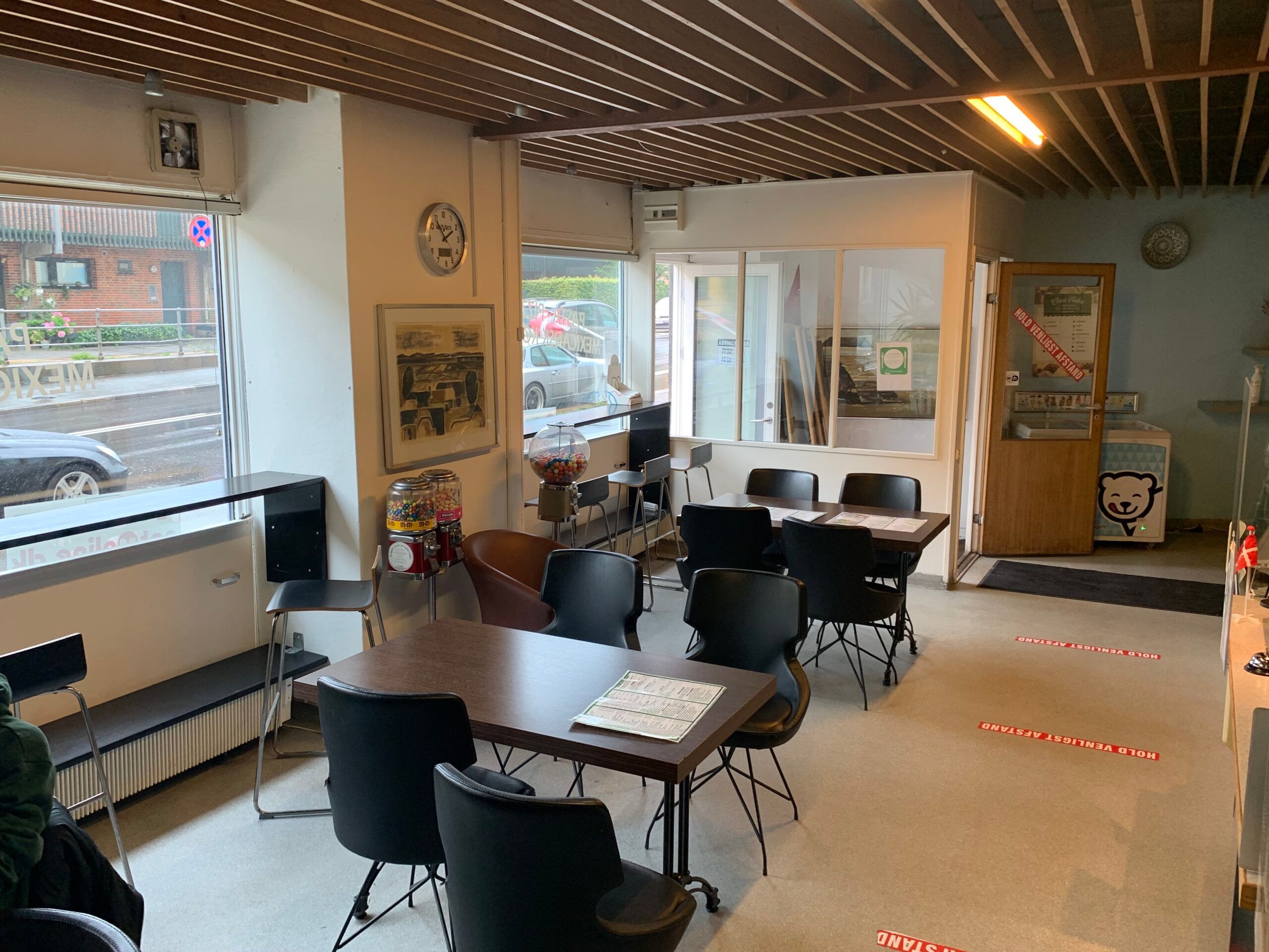 Bord og stole i lyse lokaler med vinduer. Gulv med røde mærkninger. Vægmaleri og ure. Gummiboldmaskine. Åbne glasdøre.