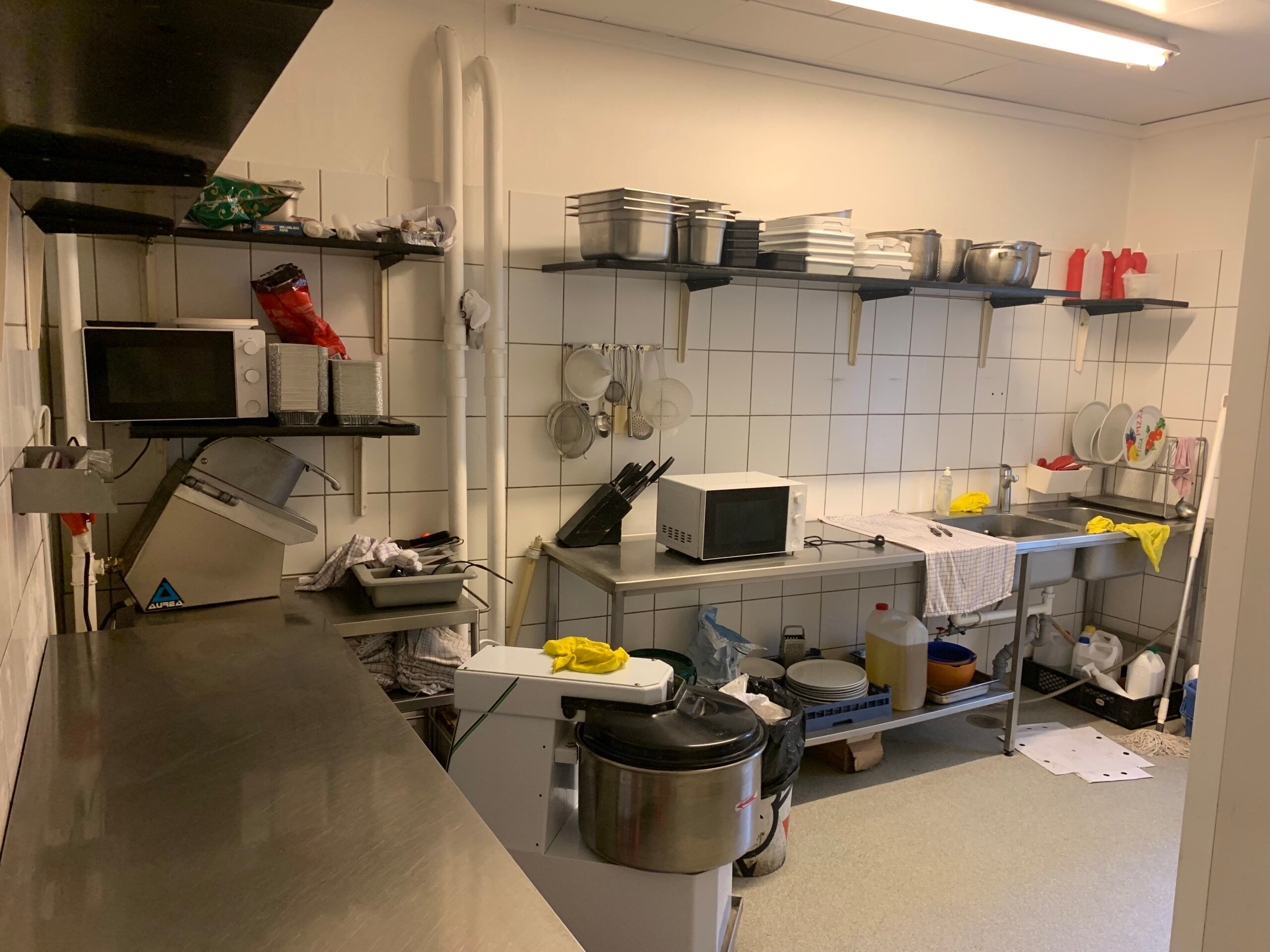 En lille køkken med to mikroovne, gryder og stålarbejdere samt en vaskemaskine, der står på en arbejdsbord. Der er et bord