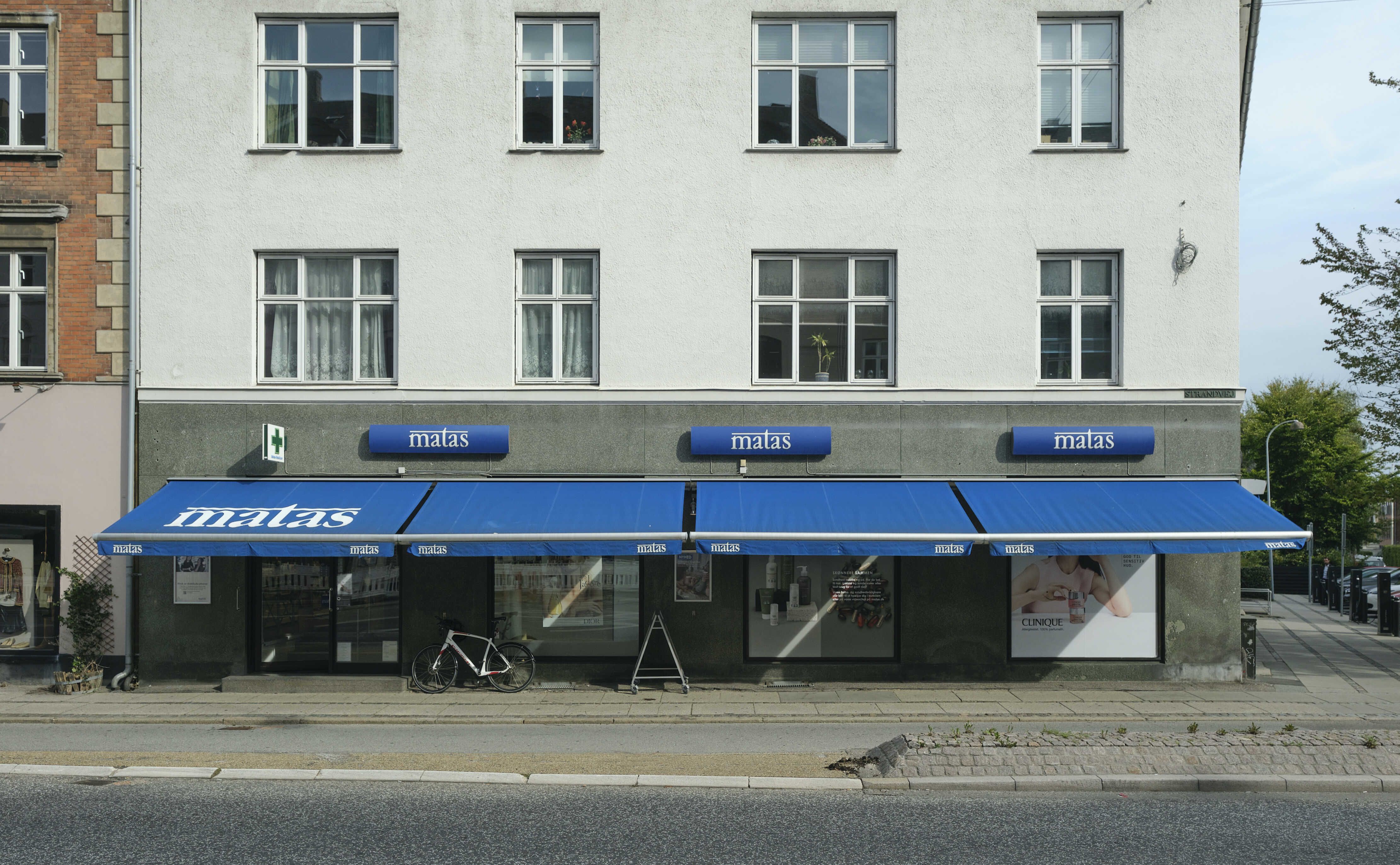 En Matas butik med en blå markise og tre skilte med Matas logoet står på Strandvejen i Hellerup, og der er en cykel parkeret