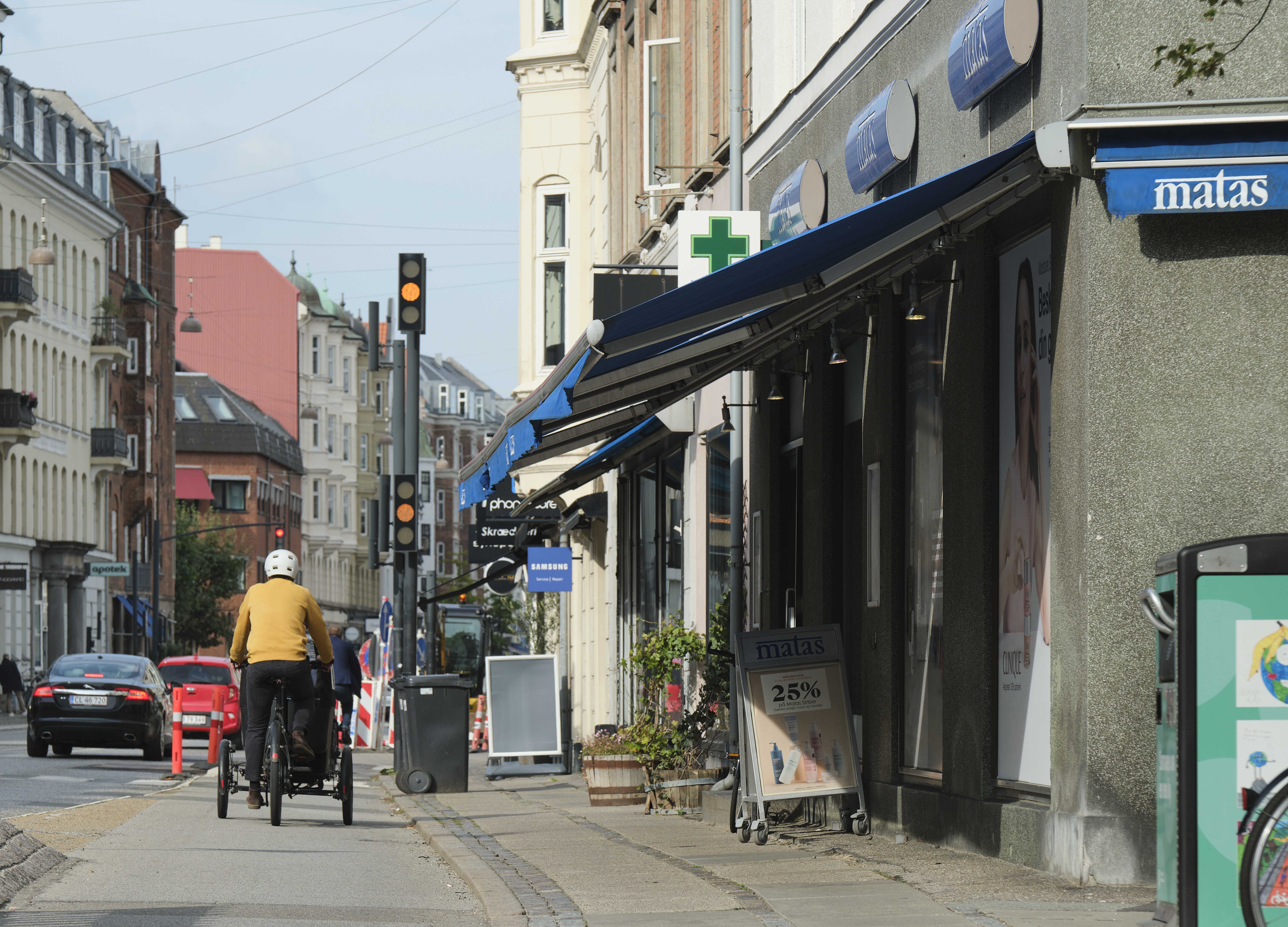 En person kører på en cykel med en vogn foran på en gågade og passerer foran et Matas-butiksvindue med en rabatskilt og en