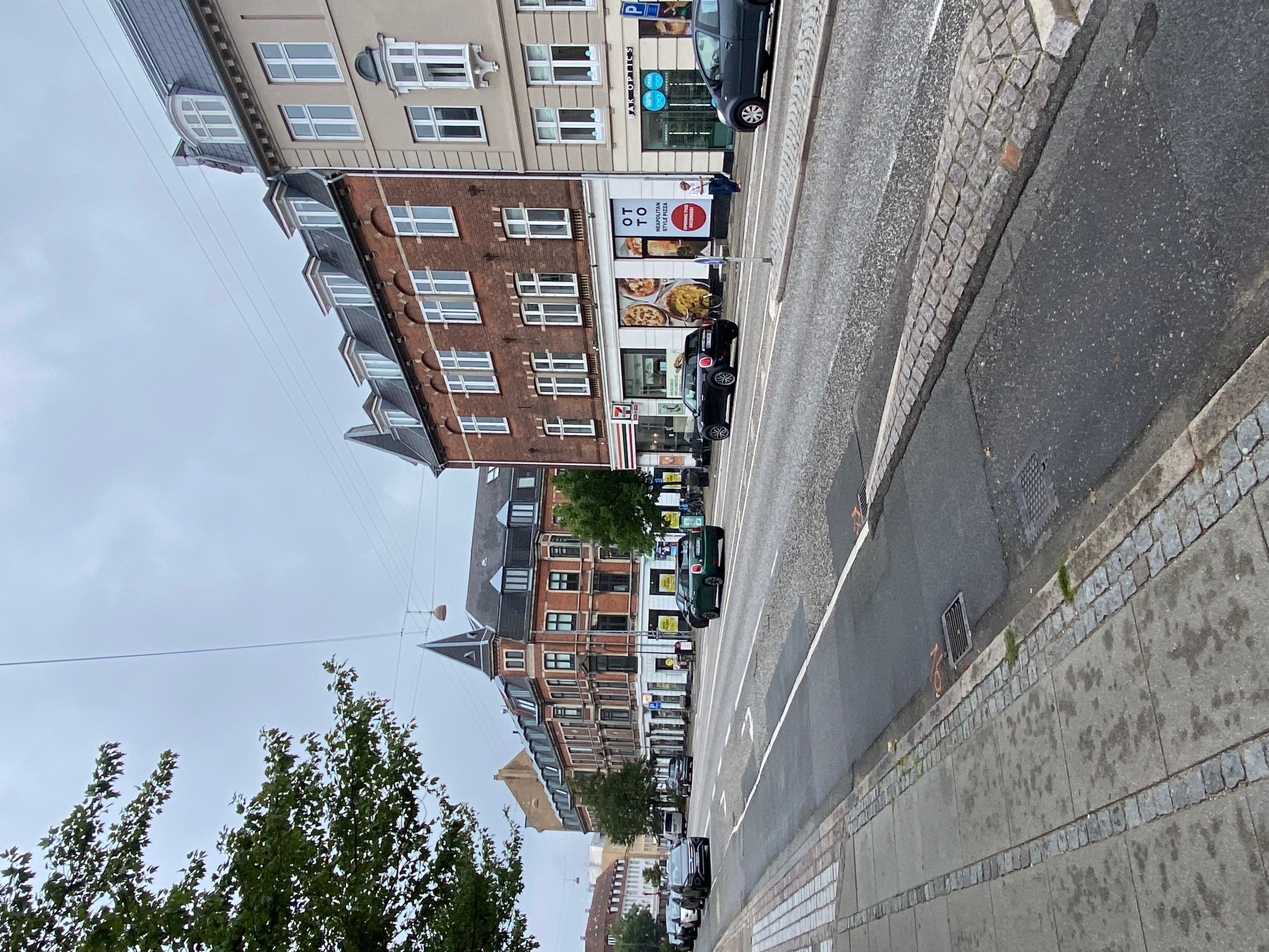 Strømledninger og et skrånende hus med reklameskilte og parkerede biler og et fortov med belægning af puds og mursten