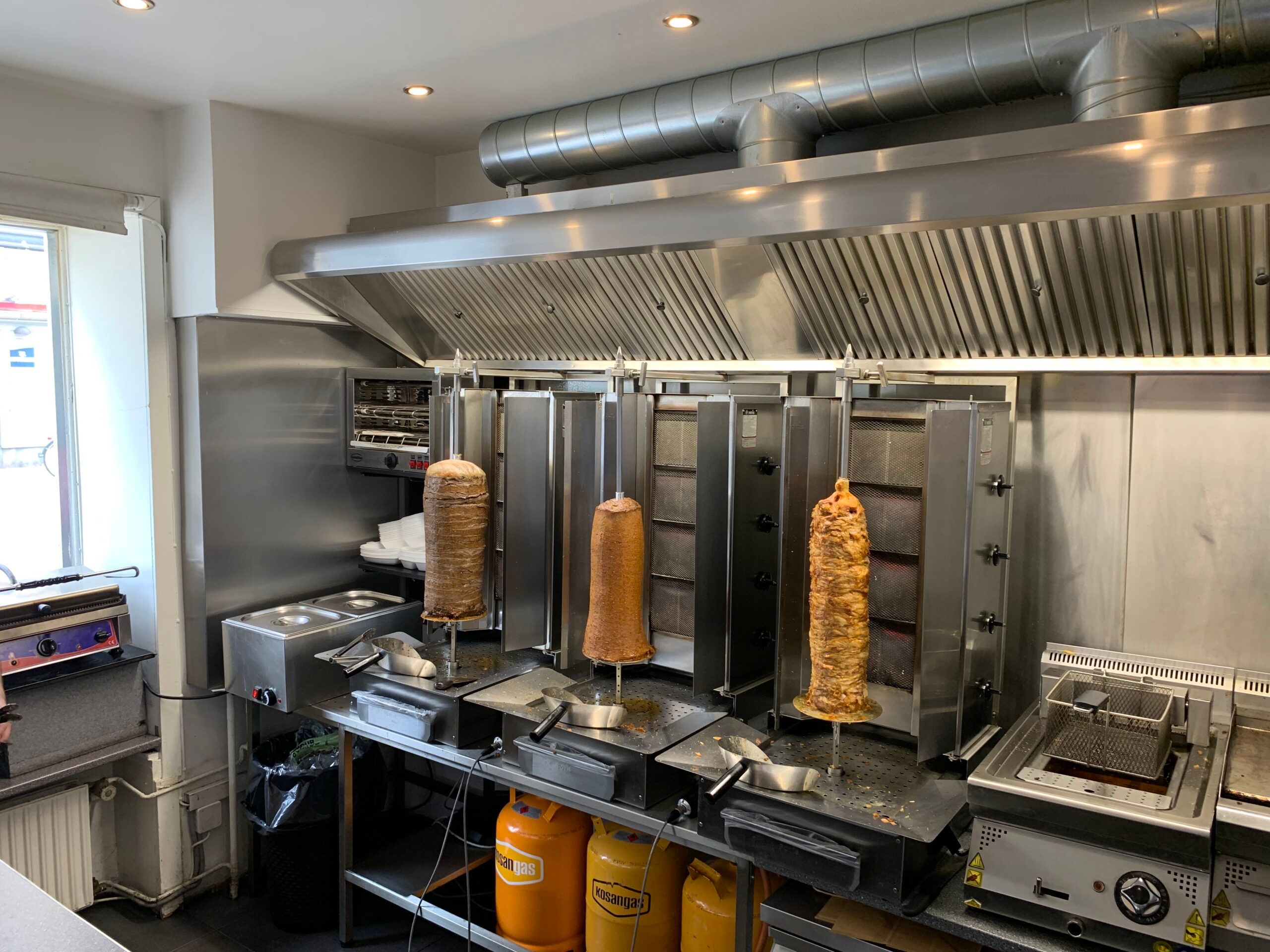 Kebab-køkken med tre spidse rotisserie-ovne og kogte rullekager og kød. Ovenfor hænger en ventileret loftslampe og der er