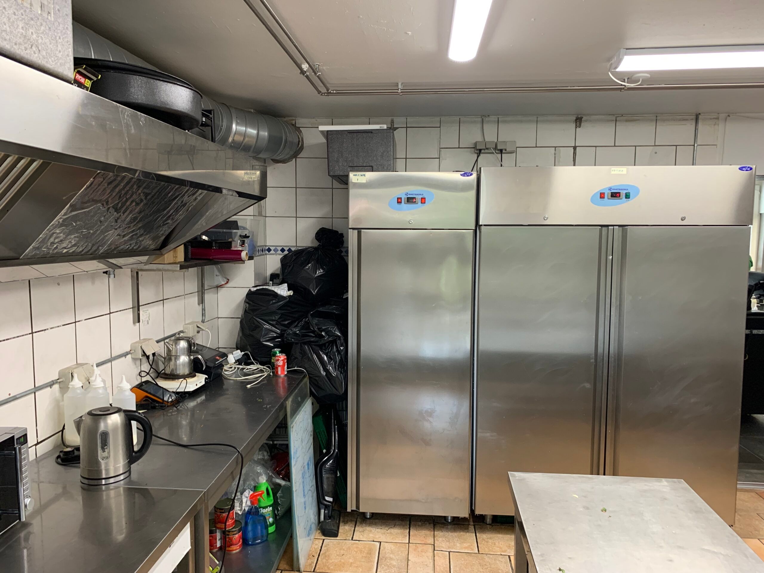 To store køleskabe står i en kommerciel køkken, der har en mikrobølgeovn, en kasse, en kaffemaskine og et skrald.