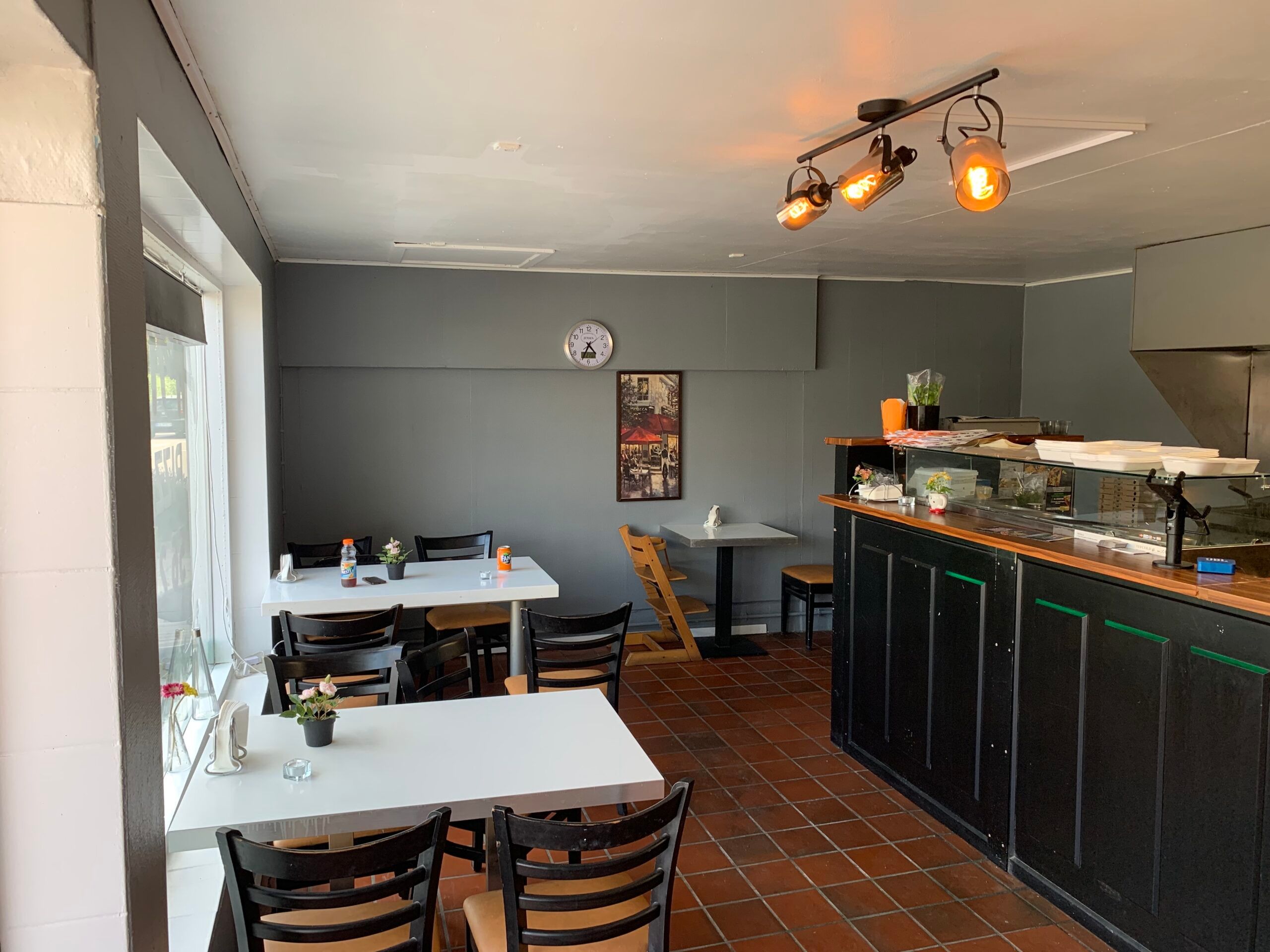 En møbleret restaurant med bord og stole, gulv med røde fliser, et disk og en simpel køkkenudstyr bag disken, og en vægur