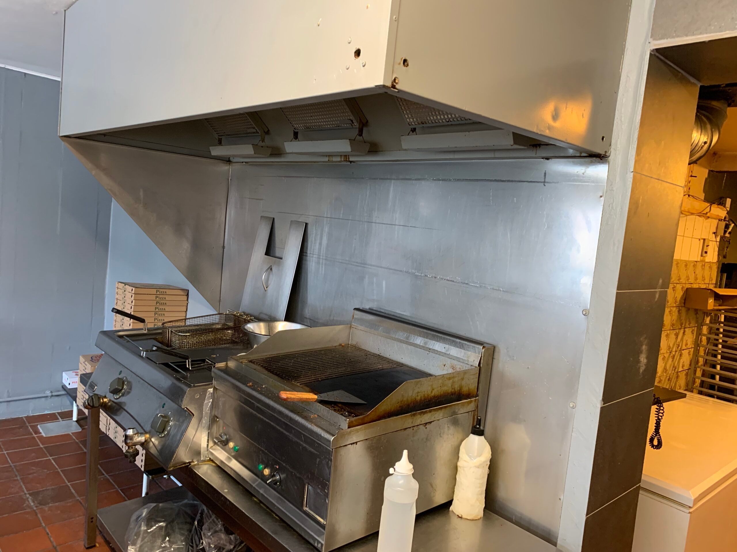 Pizzaovn med grill og røgsuger i stående position, med to flasker og en kuglepen og kasse med pizzaer i baggrunden