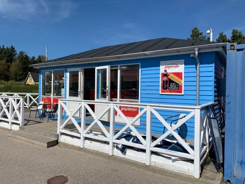 Langlejender kaffebar og kiosk med udendørs siddepladser og Coca-Cola reklame på facaden