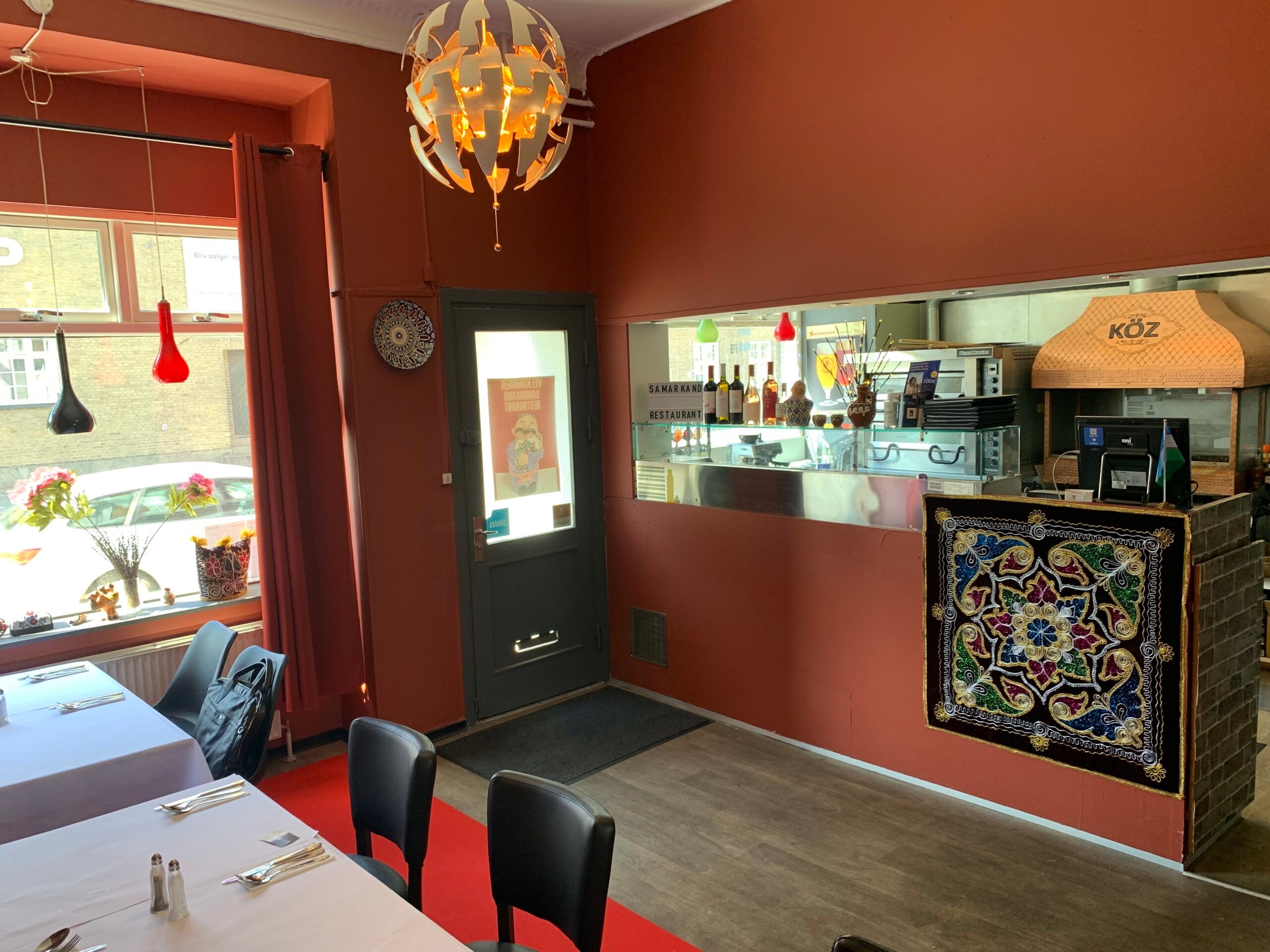 Restaurant Samarkand har flere spiseborde og stole, en indgangsdør og et lysekron. Der er en udsigt til en blomster og en
