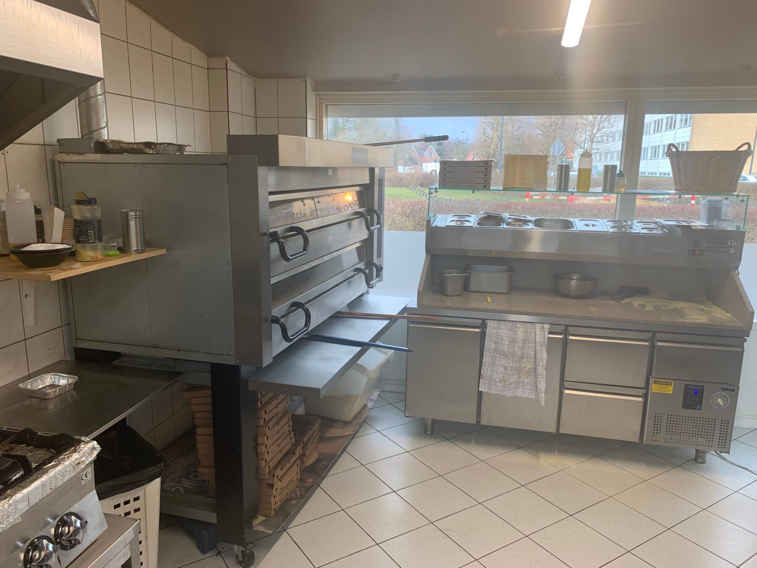 En køkken med komfur, pizzaovn, køleskabe og kogeblok med torvægge og hvide kakel.