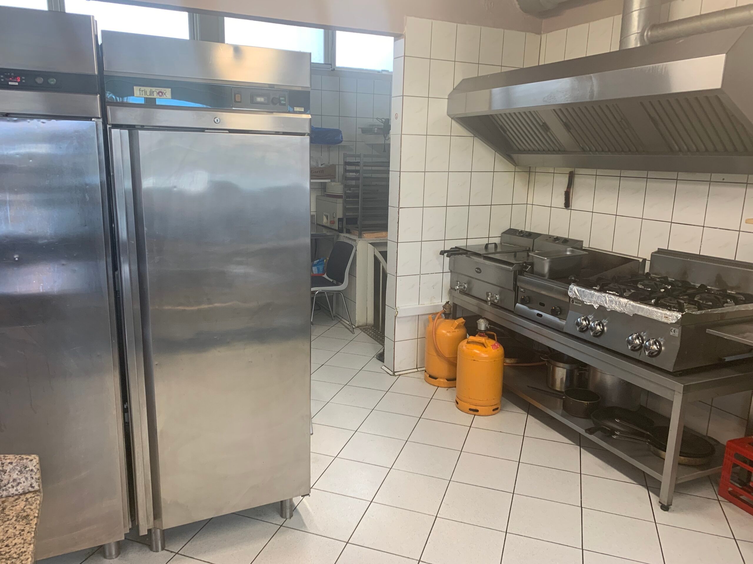 Et køkken med store køleskabe, gaskomfur og røgaffugter samt gulv og vægge i hvide kakel.