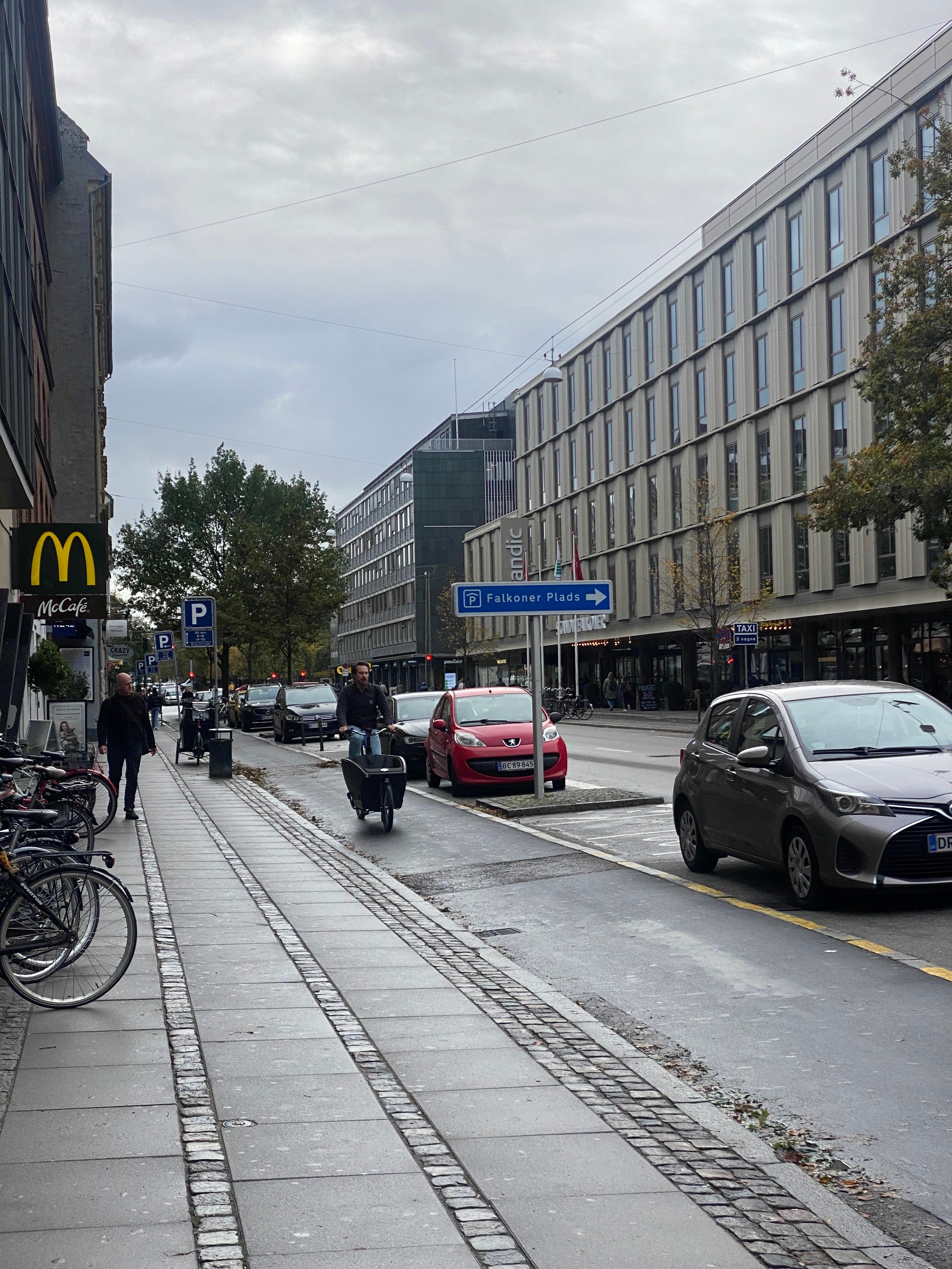En cykelrytter kører langs en gågade med en cykel med en vogn, og foran ham er en parkeringsplads med parkerede biler.