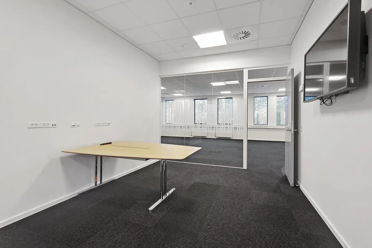 Minimalistisk kontor med hvidt loft og vægge, bord med ben i stål, tv-skærm på væggen, og åbne ruder med udsigt til træer