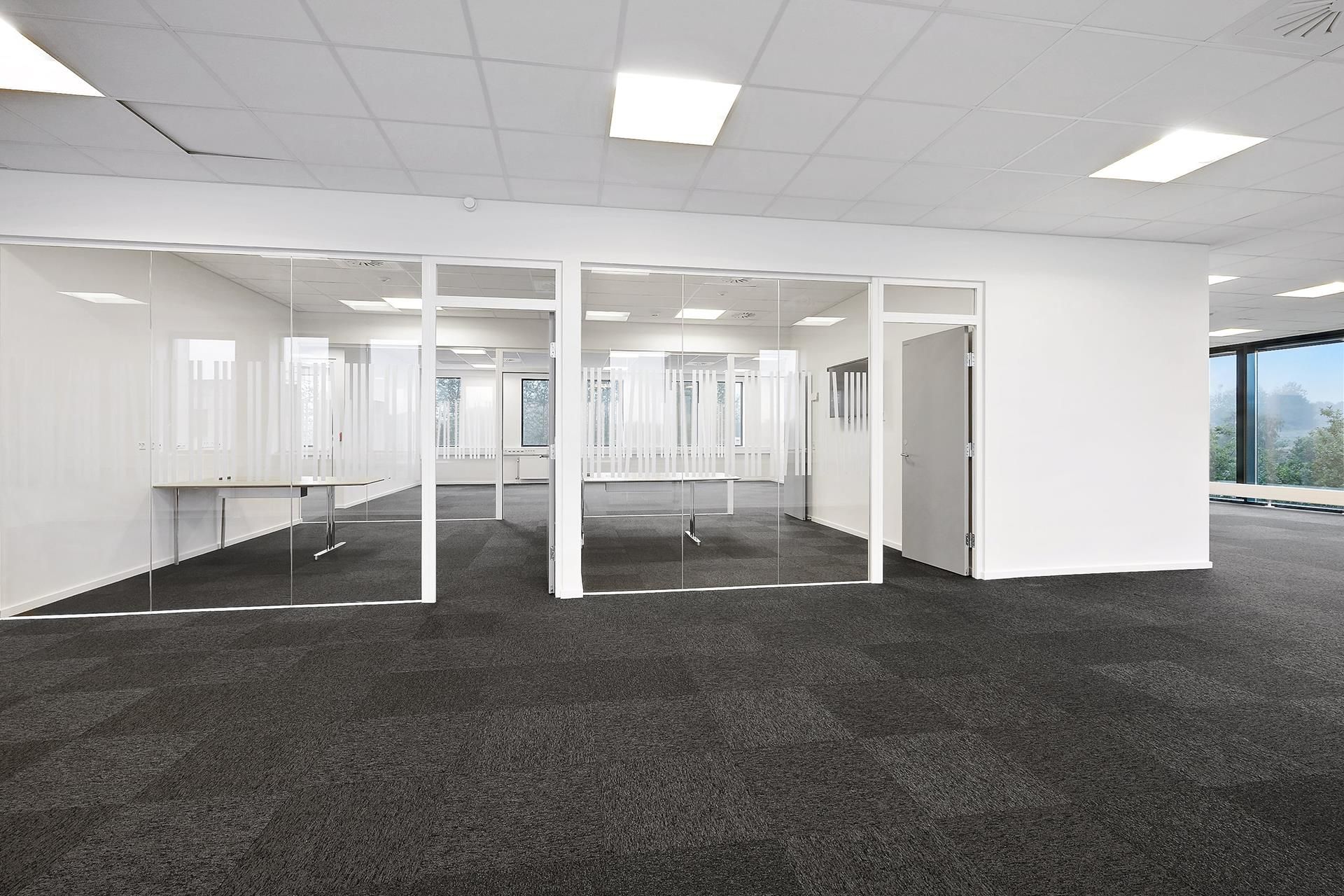 To store glasvægge og et åbent kontor med et bord og et skrivebord i en rummelig kontor. Væggene er hvide, og der er