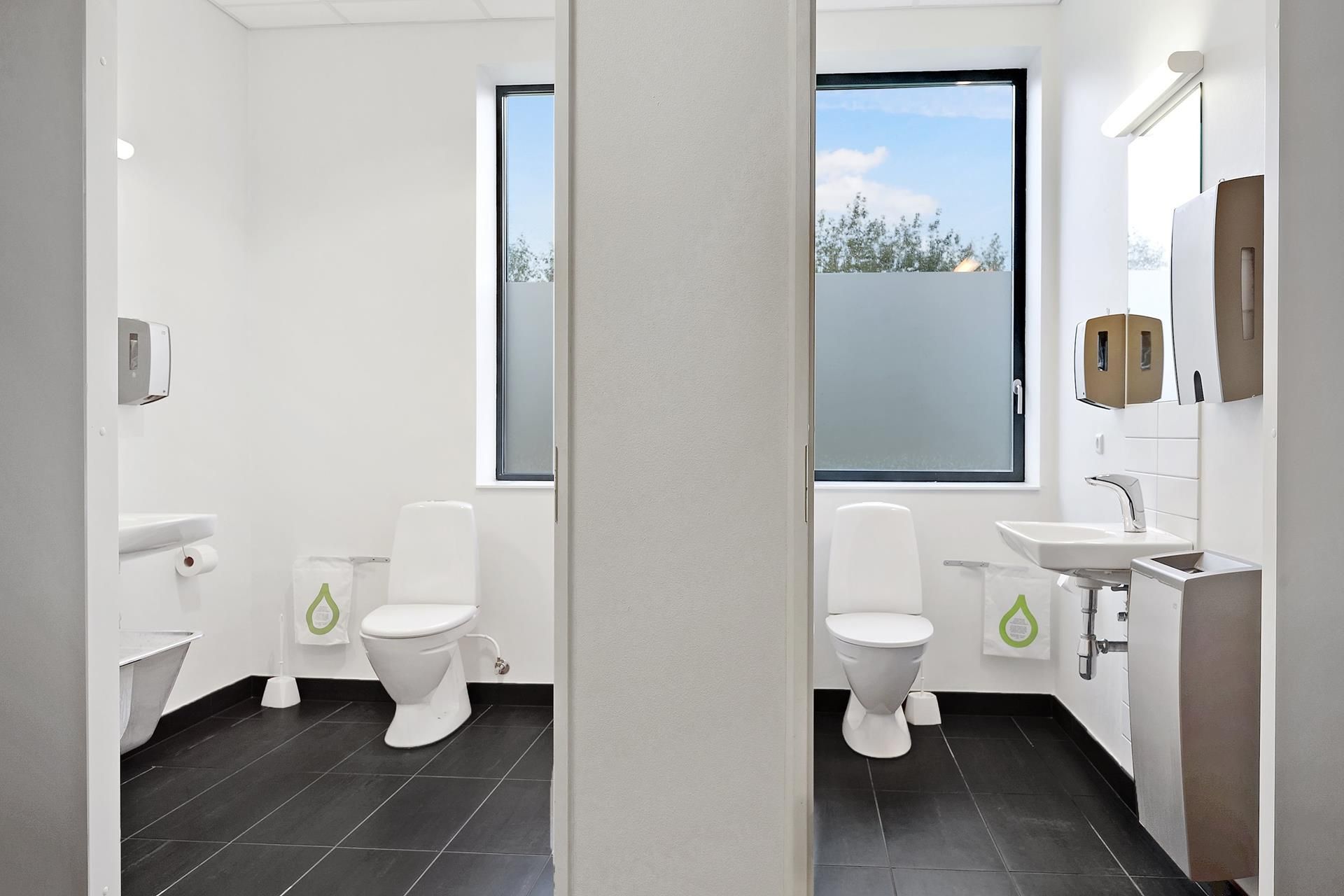 Moderne toiletrum med to toiletter og en håndvask, hvor der er tørklæder og håndsæbeautomater monteret på væggen