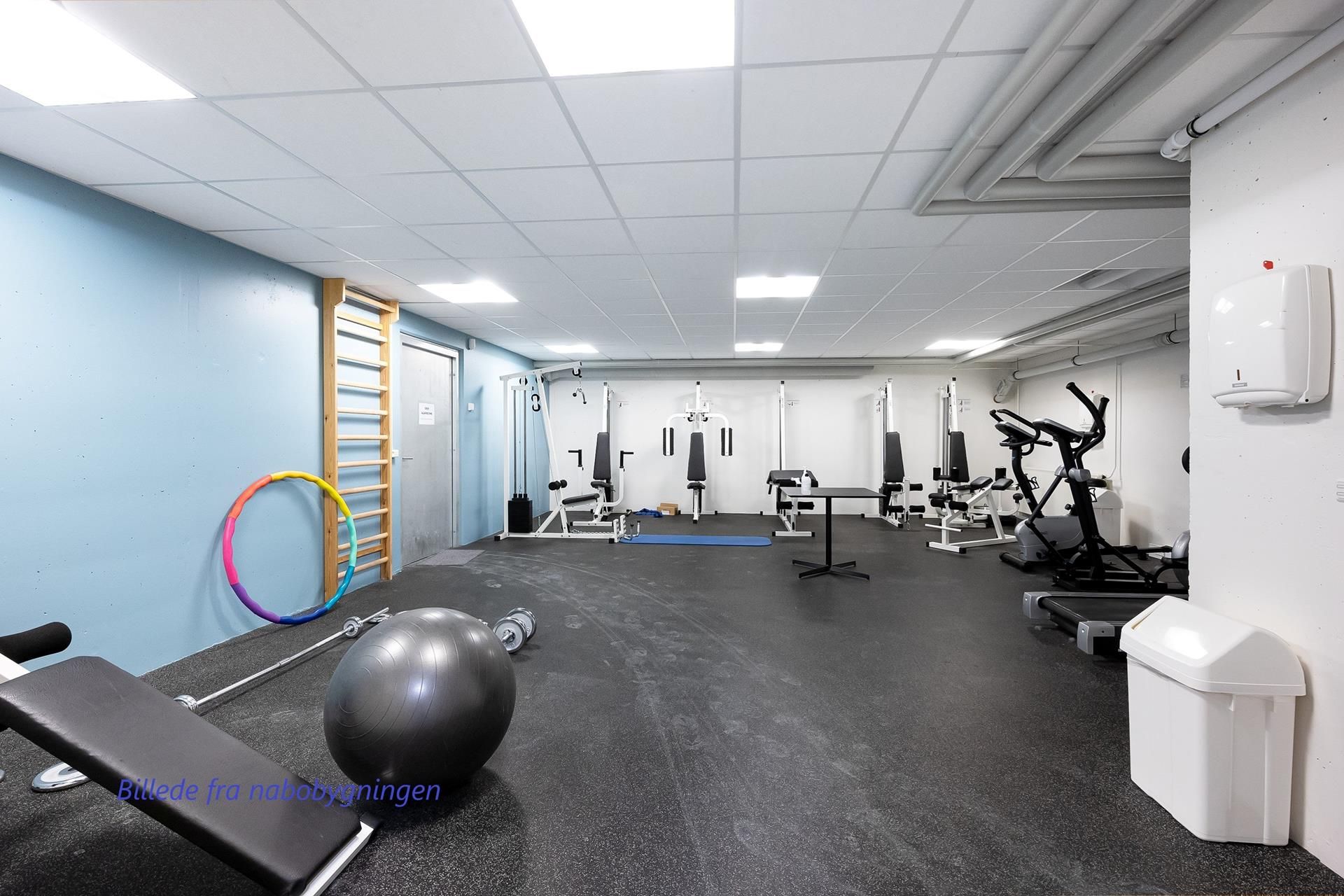 Et fitnessrum med udstyr som træningsmaskine, trappe, apparater og hula hoop. Højt lys og en ren gulvoverflade er tydelige.