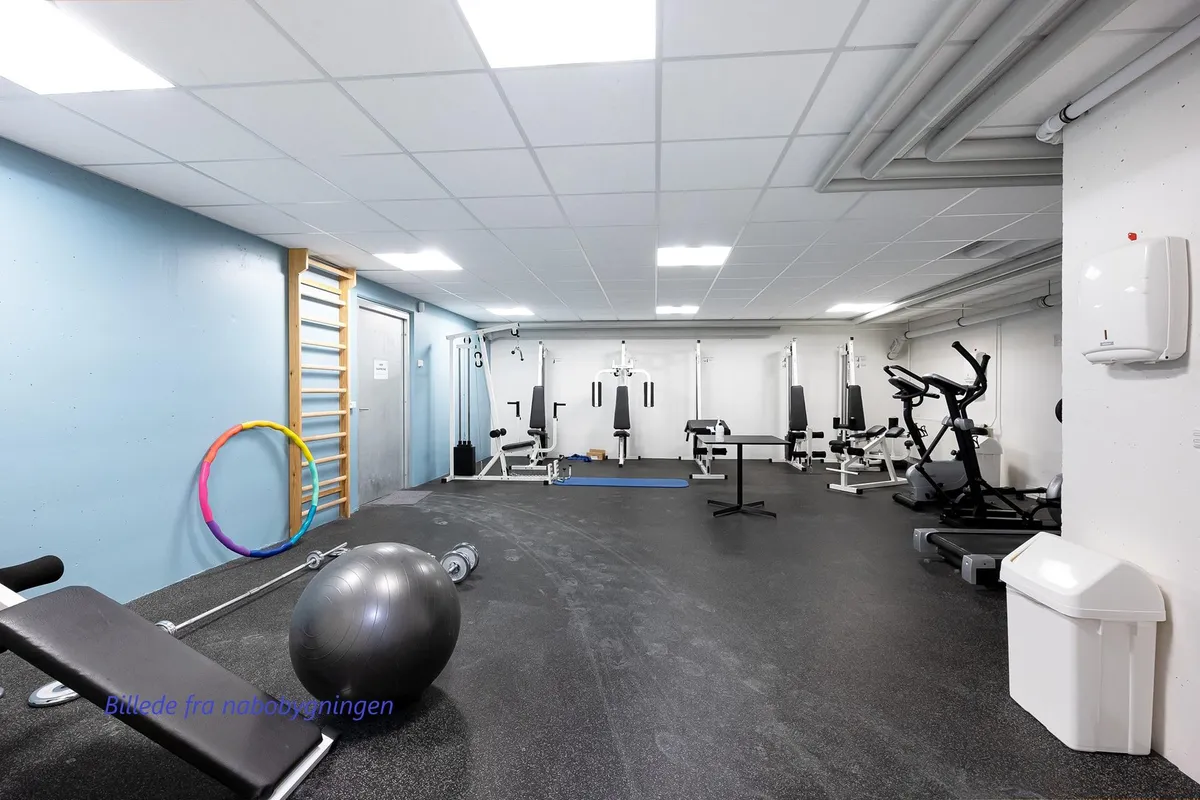 Et fitnessrum med udstyr som træningsmaskine, trappe, apparater og hula hoop. Højt lys og en ren gulvoverflade er tydelige.